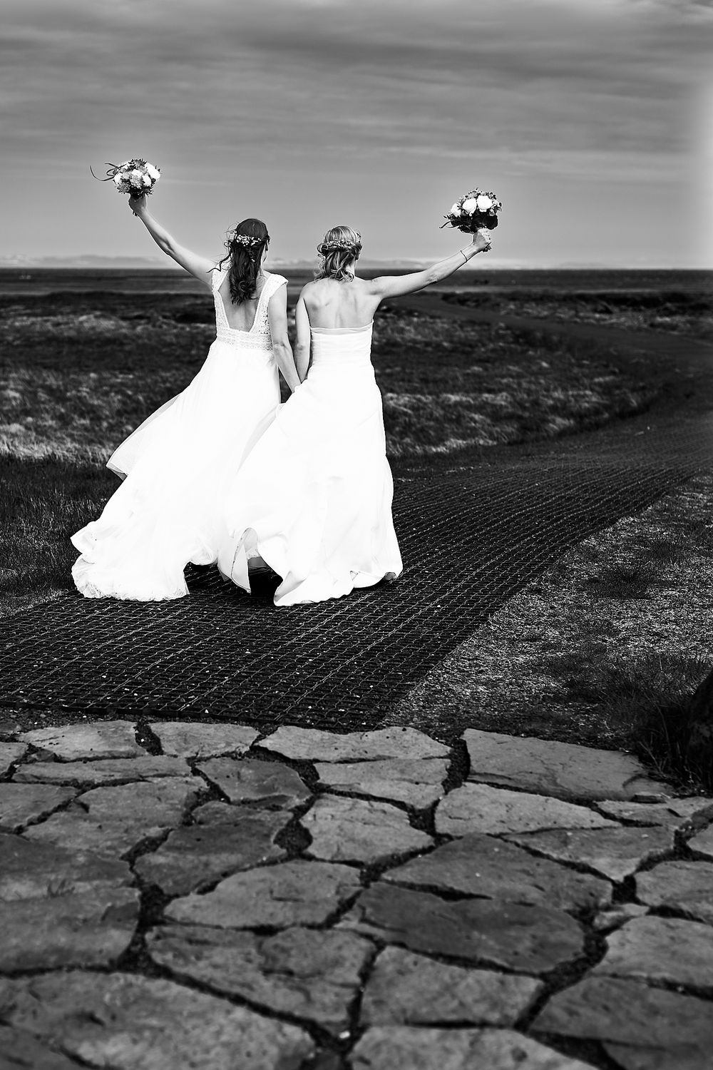 Two figures in white wedding dresses hold bouquets triumphantly on a rocky path in black and white.