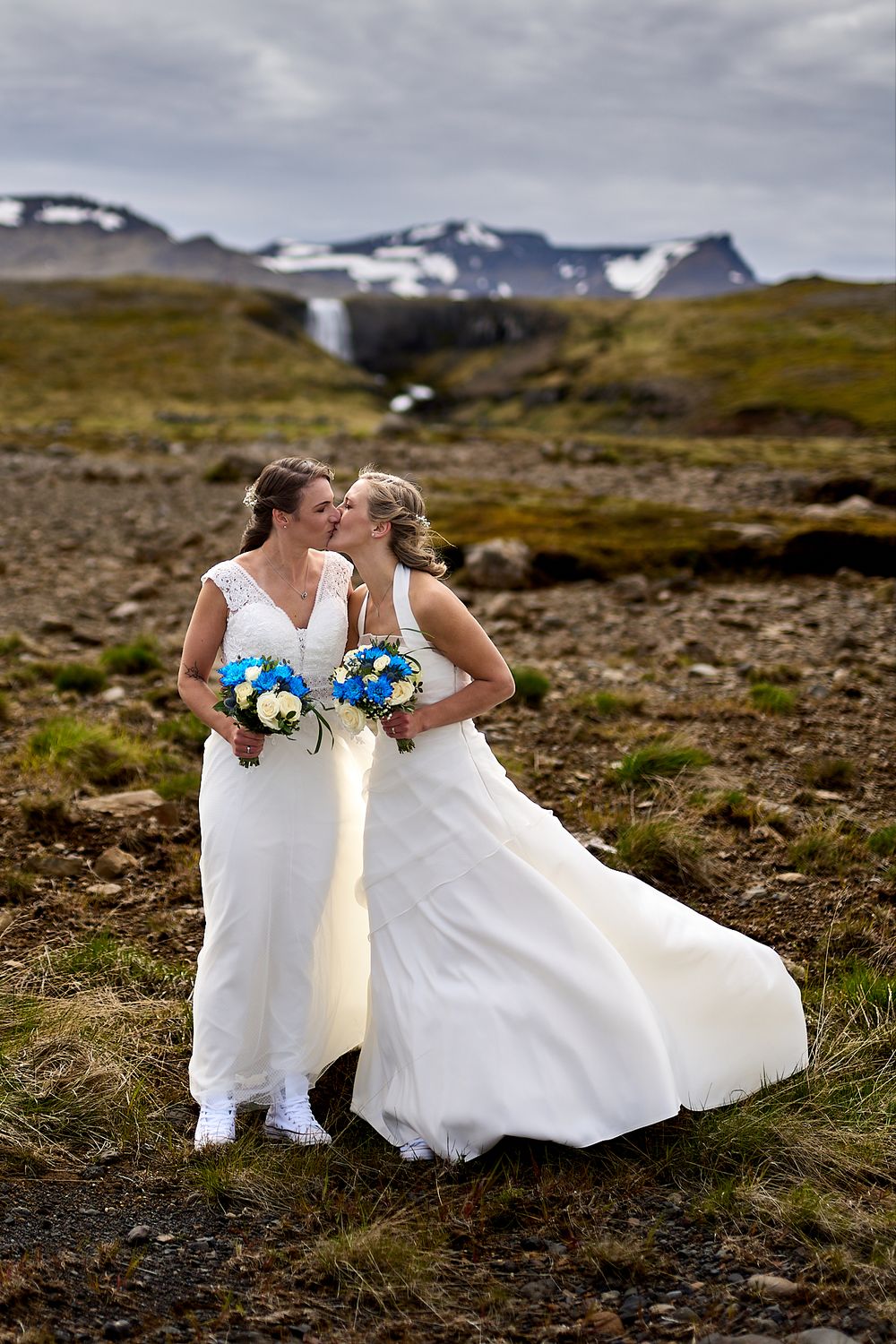 Two people in wedding dresses share an intimate moment while holding blue and white bouquets in a mountain setting.