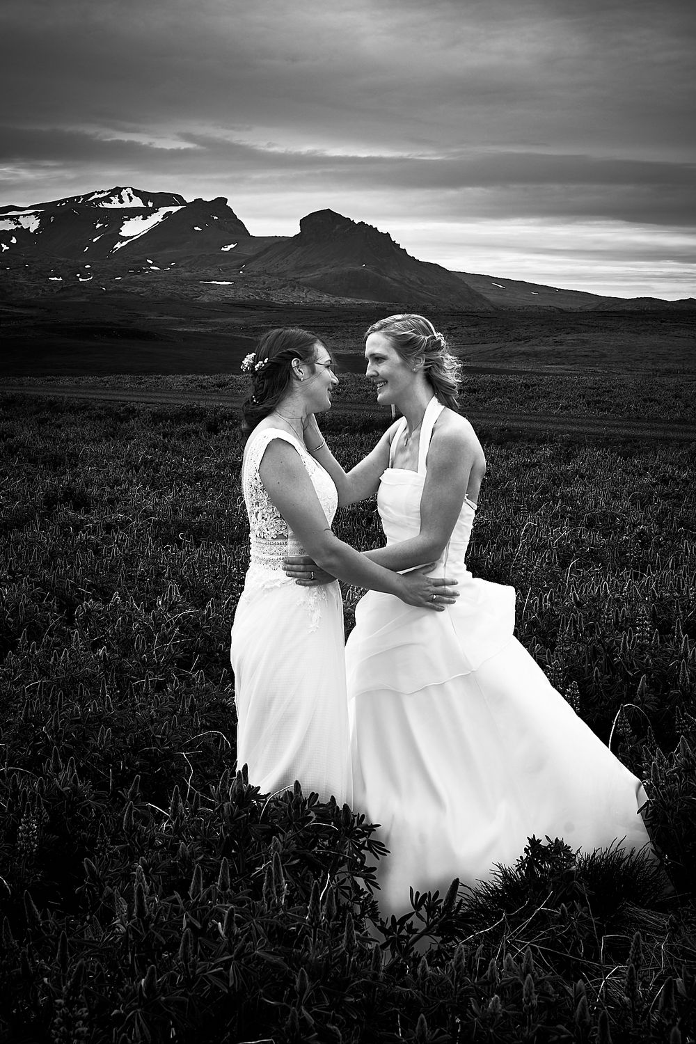 A romantic wedding portrait in black and white shows a couple in white dresses against a mountainous backdrop.