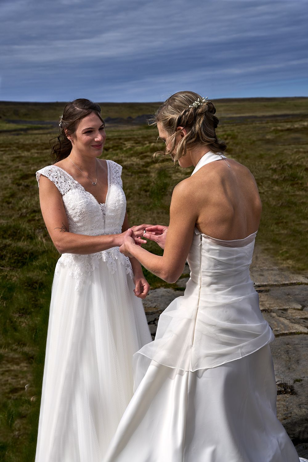 Two people in white wedding gowns share a joyful moment during a ring exchange ceremony outdoors.