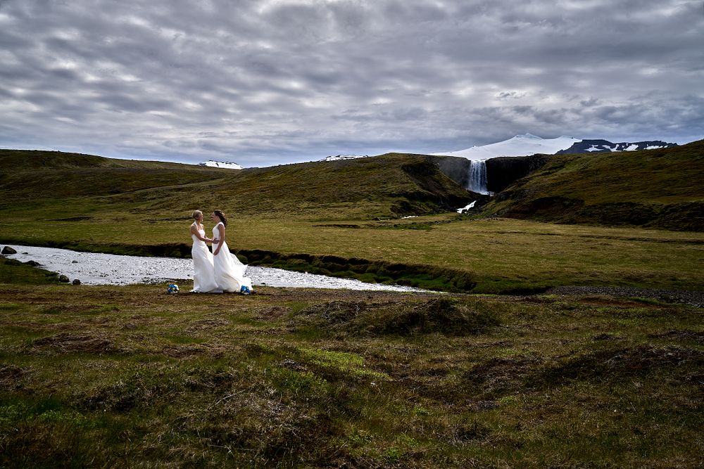 Two wedding figures pose beside a stream in a dramatic mountain landscape under cloudy skies.