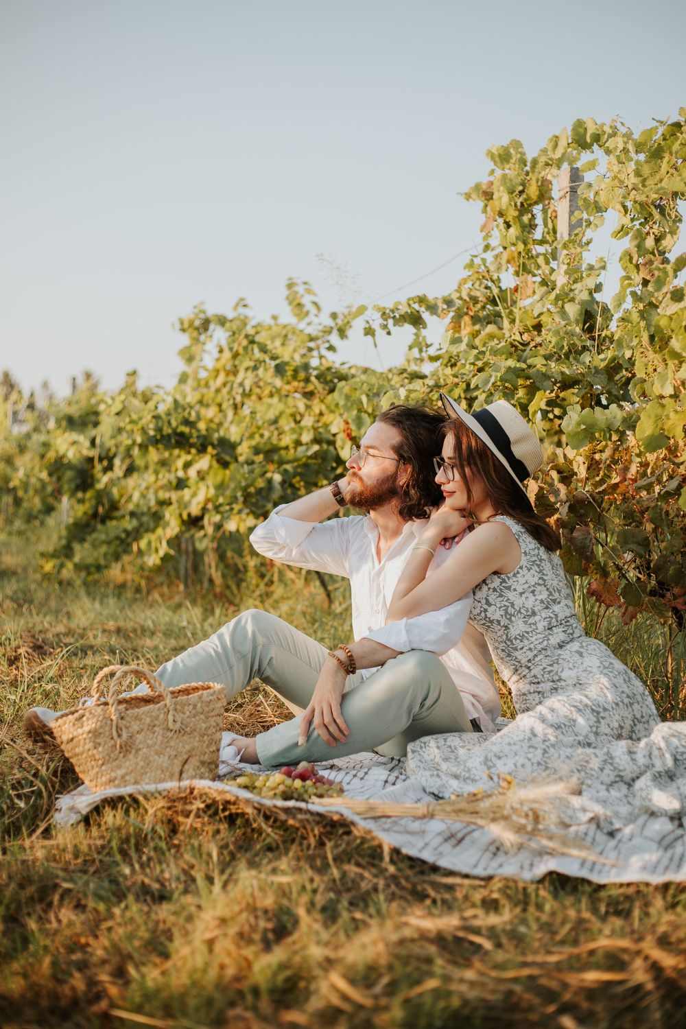 A couple enjoys a romantic sunset picnic in a vineyard with a blanket and basket.