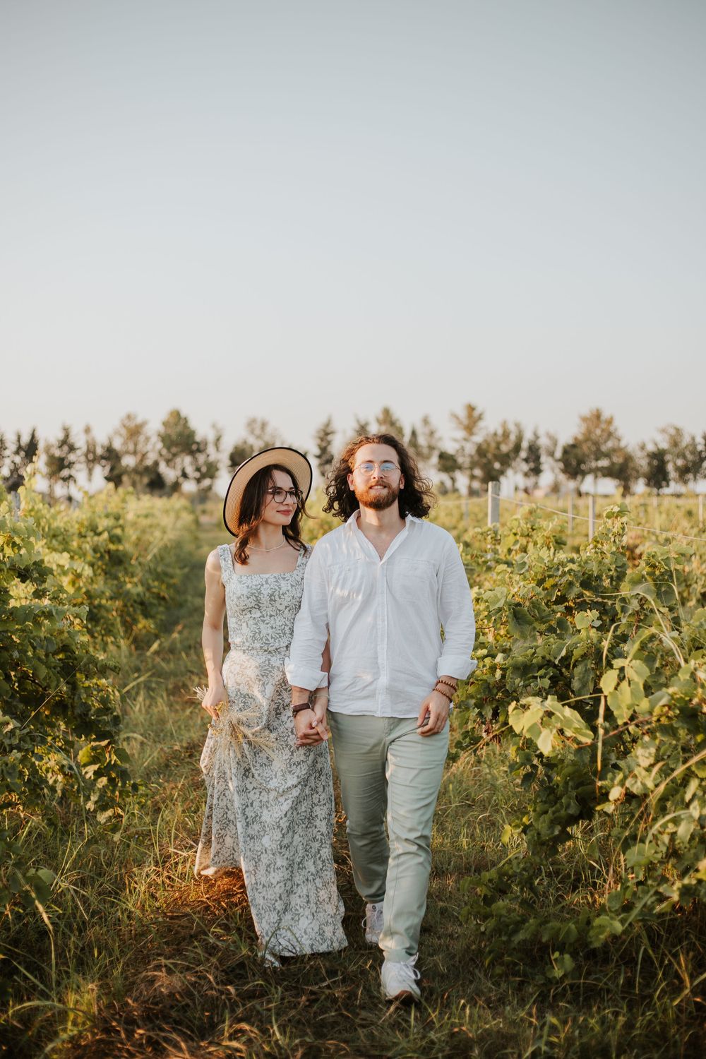 A couple holds hands while walking through a vineyard during golden hour photography session.