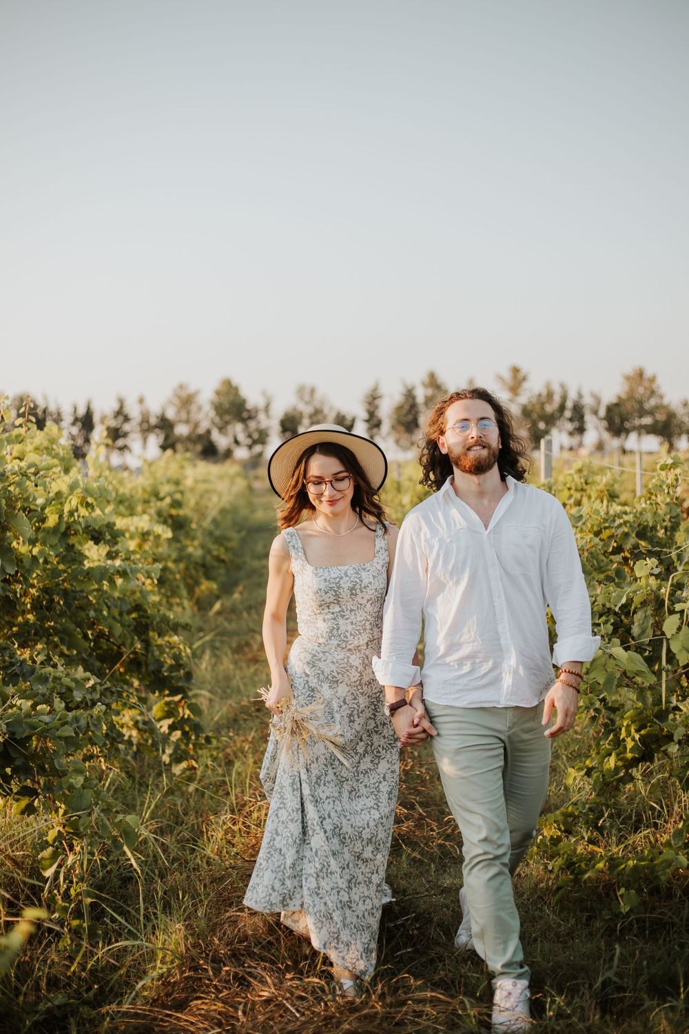 A couple smiles while walking together through a sunny vineyard landscape.