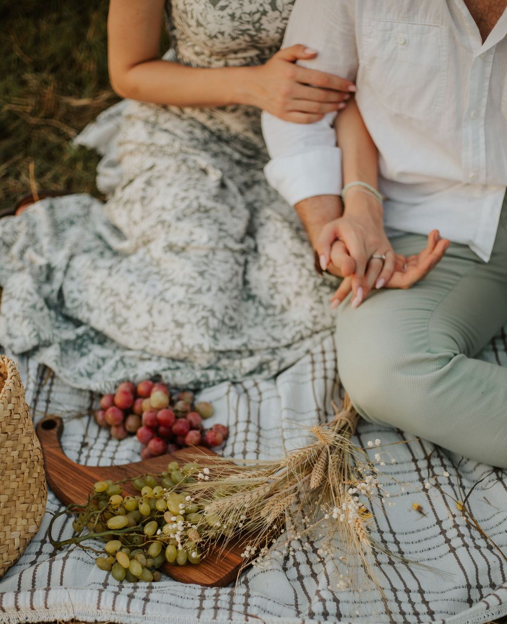 Close-up of hands and picnic spread with grapes, wheat stalks and fresh fruit on checkered blanket.