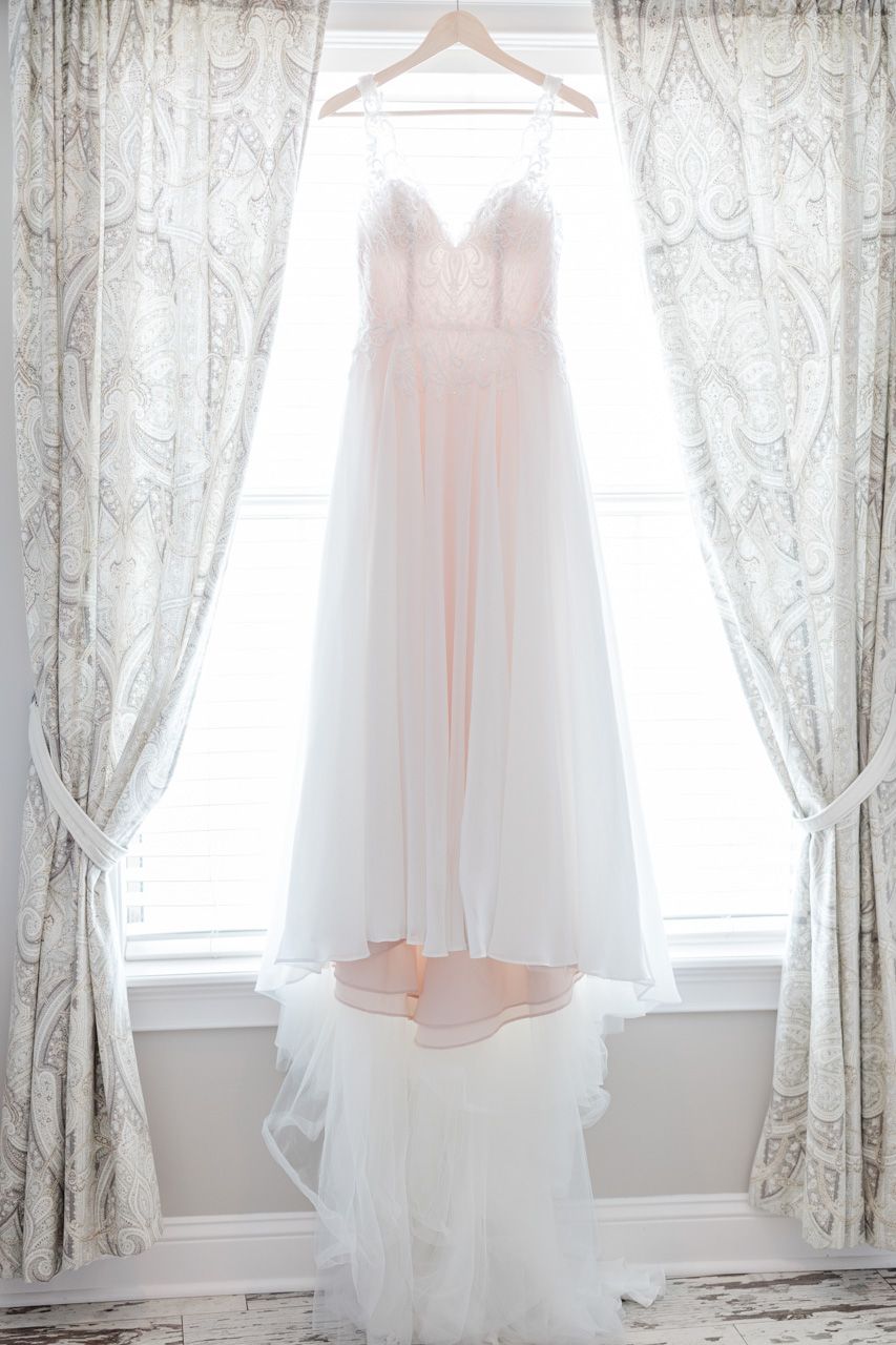 White wedding dress hanging in window with lace curtains creating soft natural lighting.