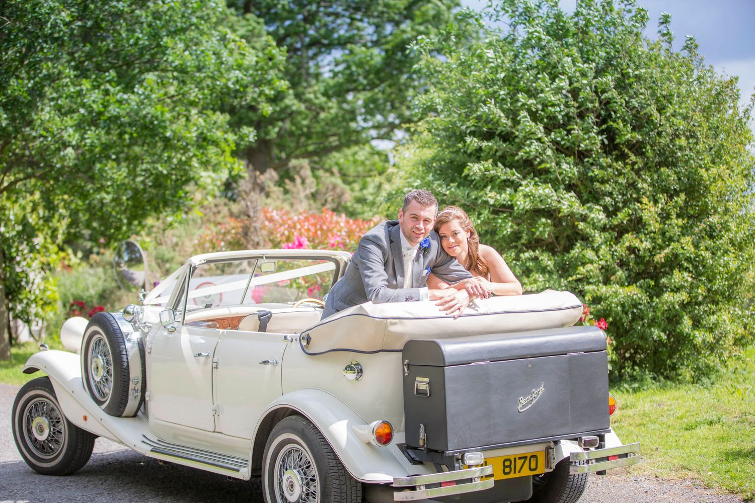A couple posing in a vintage white convertible car surrounded by lush garden scenery.