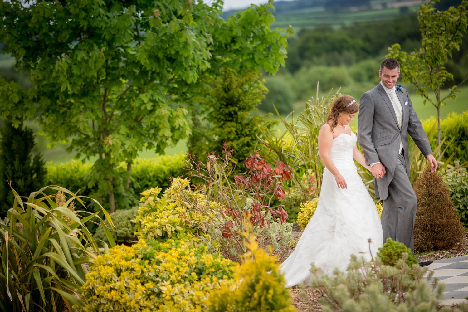 A wedding couple poses in a lush garden filled with vibrant yellow flowers and greenery on a sunny day.