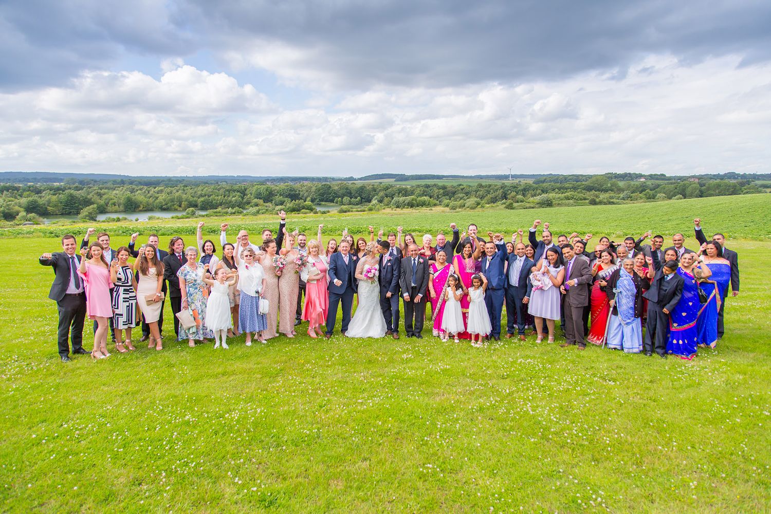 Large wedding group photo with colorful attire on scenic hilltop meadow.