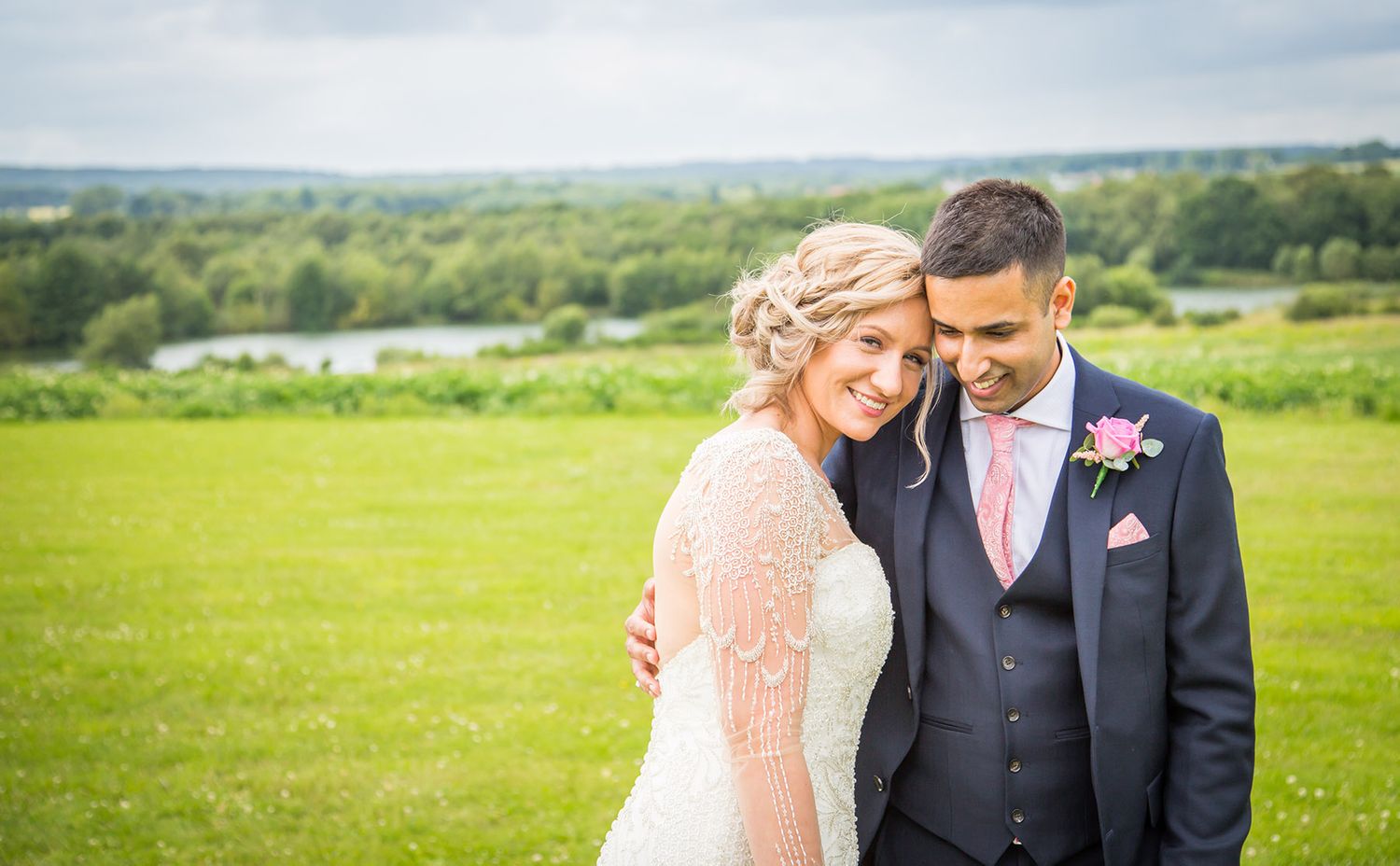 A romantic outdoor wedding photo showing a couple embracing in a green countryside setting.