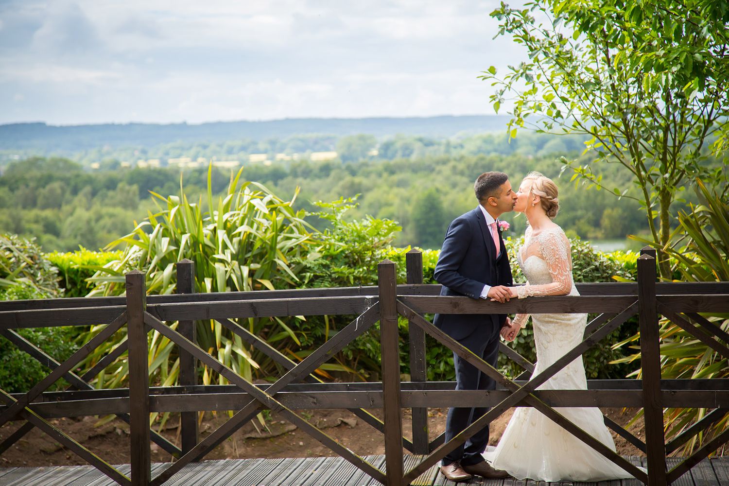 A wedding couple sharing an intimate moment on a wooden bridge overlooking a scenic landscape.