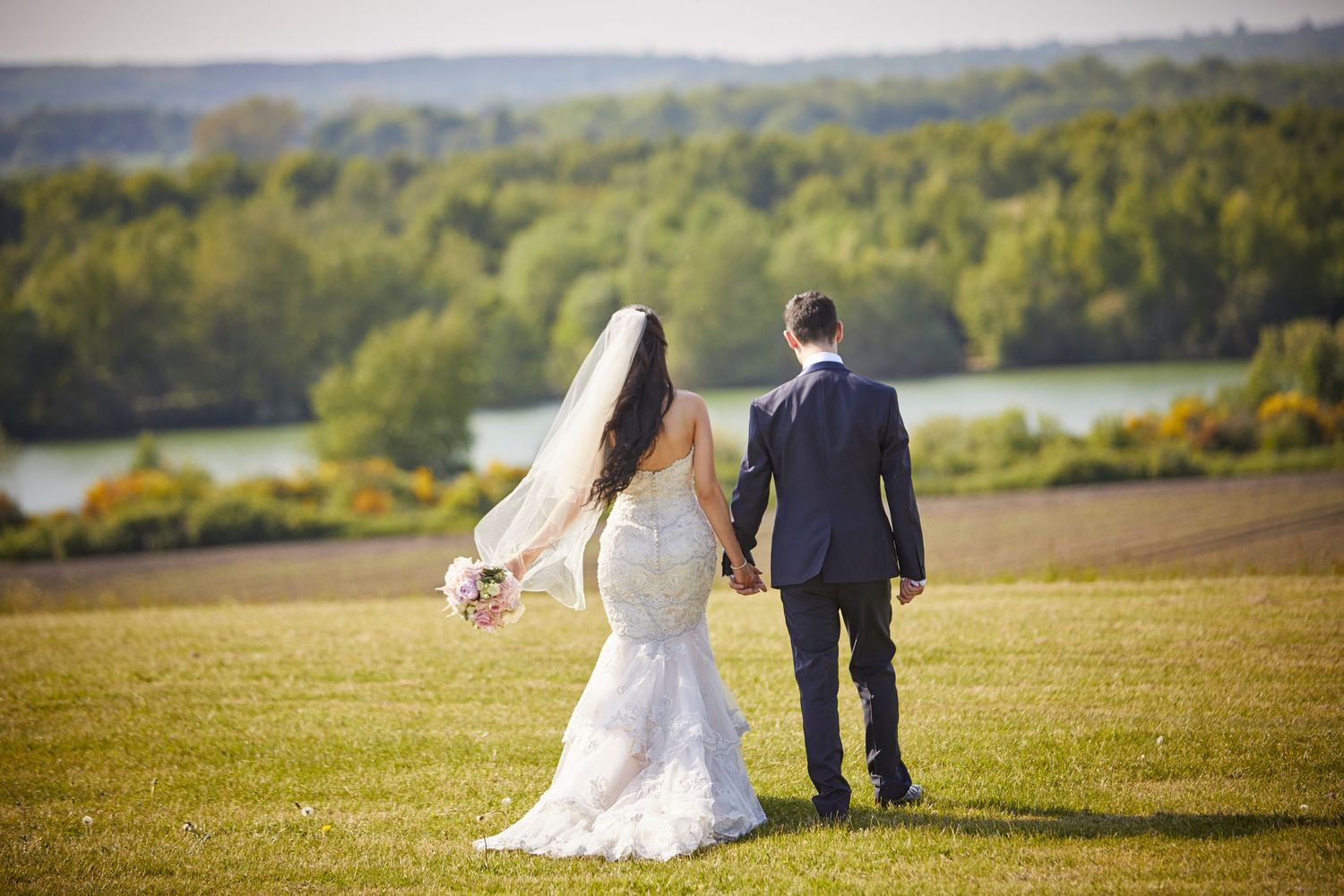 Newlyweds walk hand in hand across a hilltop meadow overlooking a scenic lake.