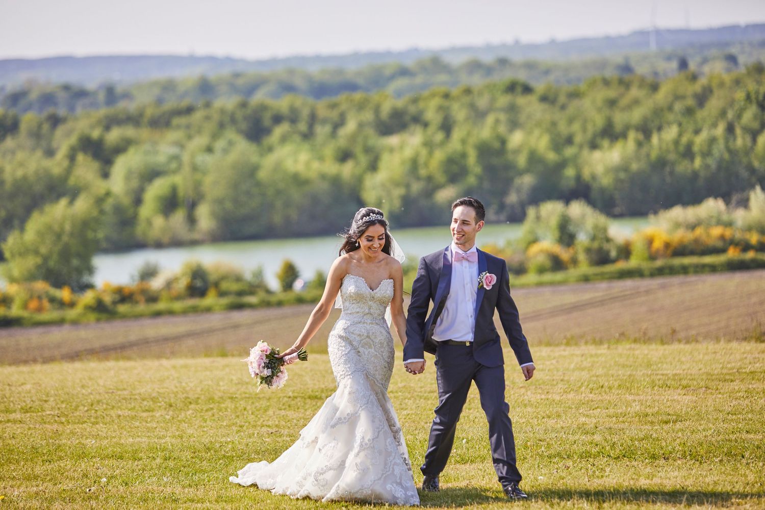 Newlyweds walk joyfully across a hilltop field with a lake in the background.