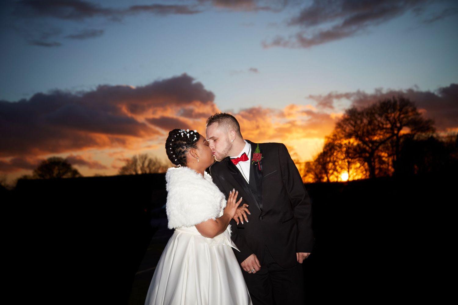 A dramatic sunset silhouettes a couple in wedding attire against an orange and purple sky.