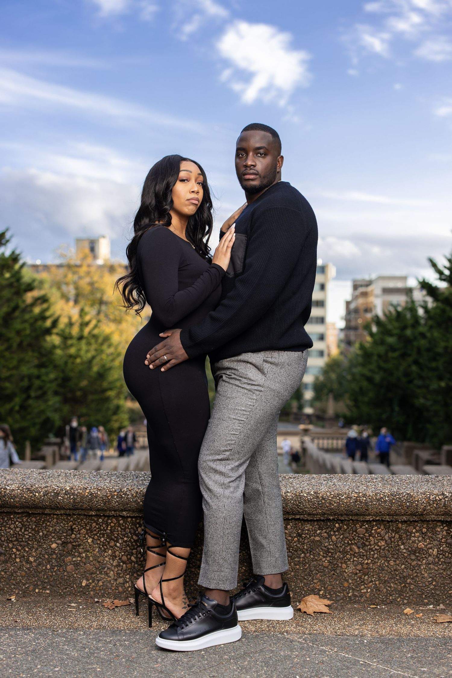 Couple in coordinated black outfits embrace on stone wall with city buildings in background.
