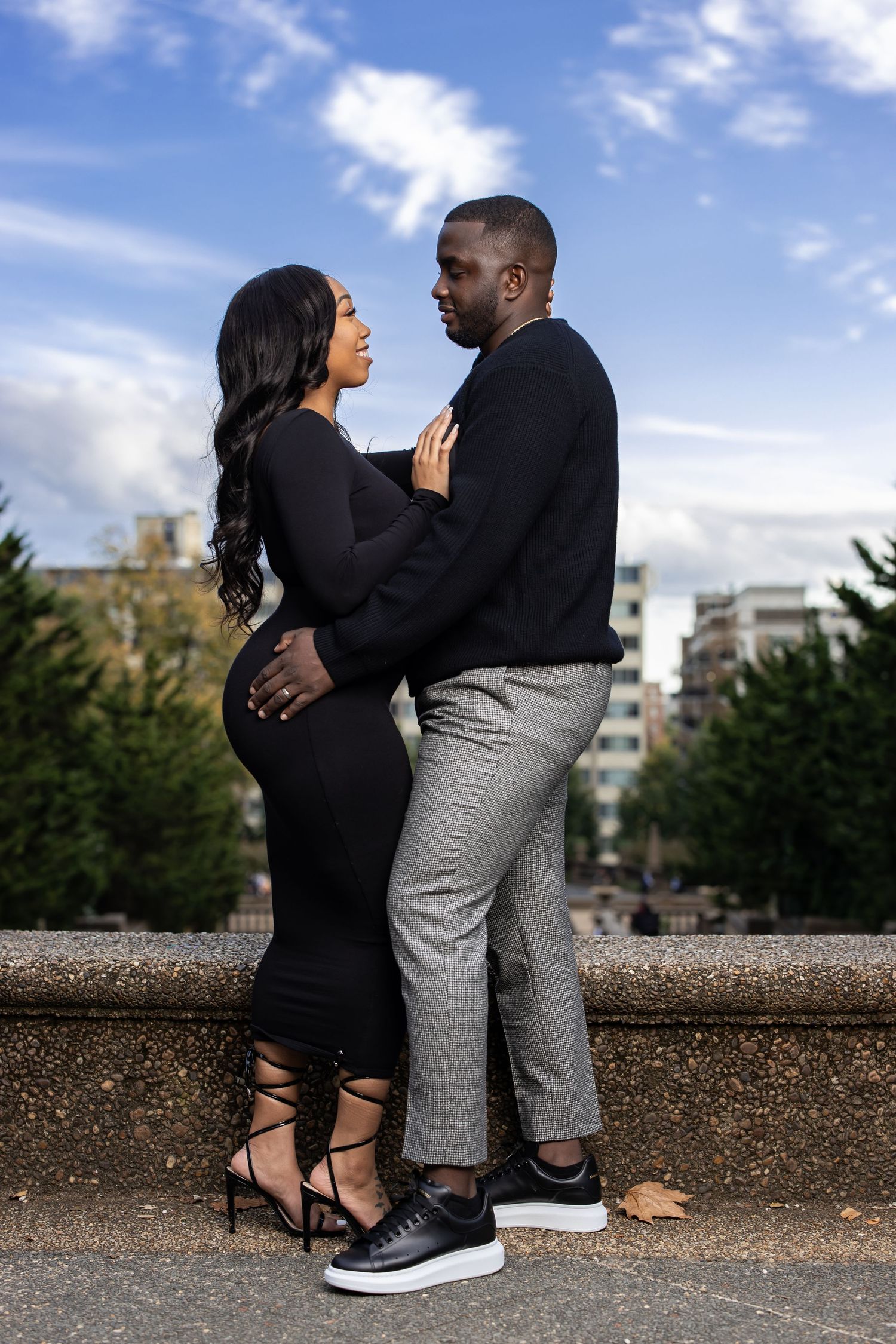 Couple in black outfits embrace romantically against blue sky and urban backdrop.