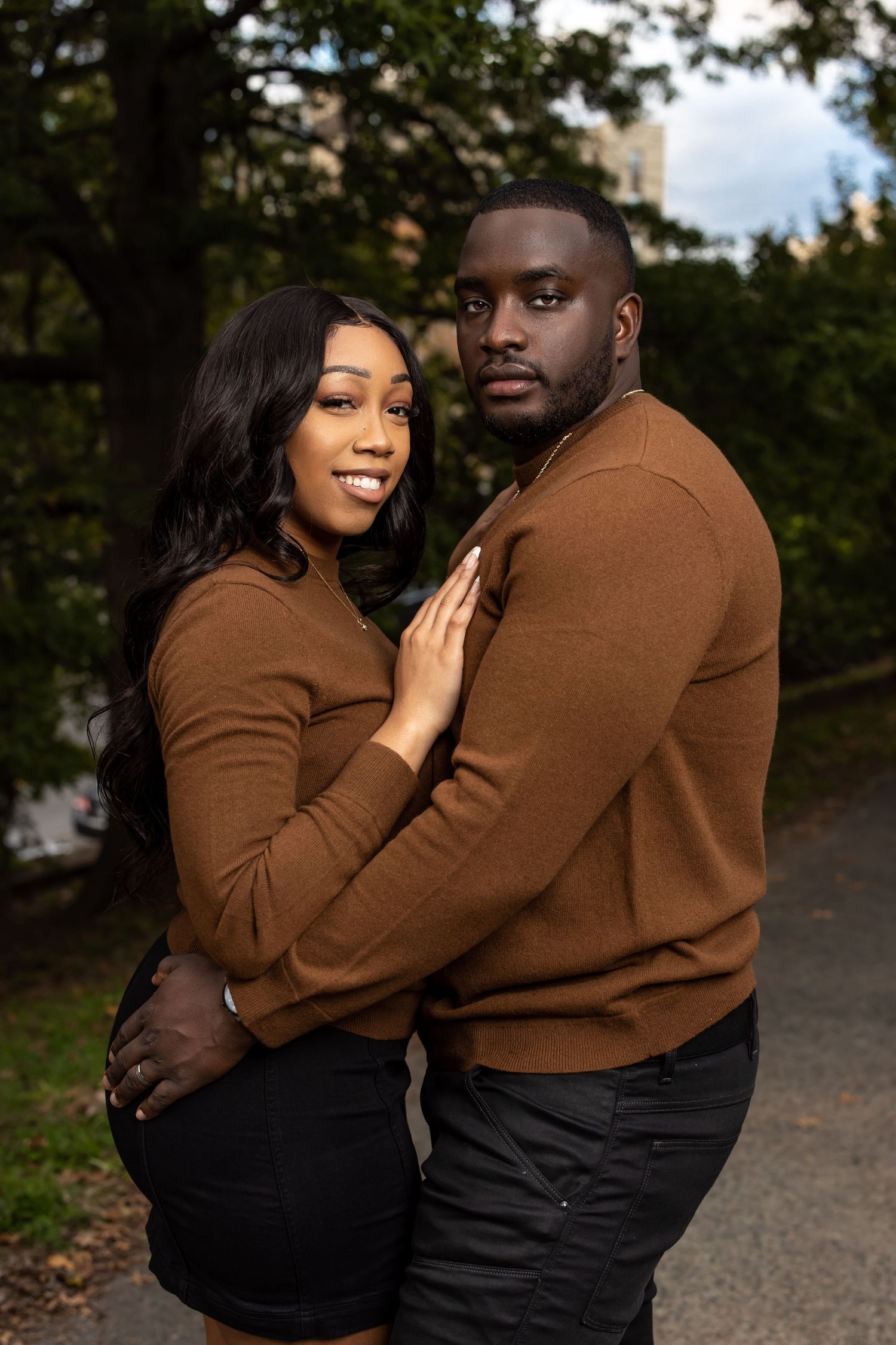 A romantic couple in matching brown sweaters embrace outdoors on an autumn path.