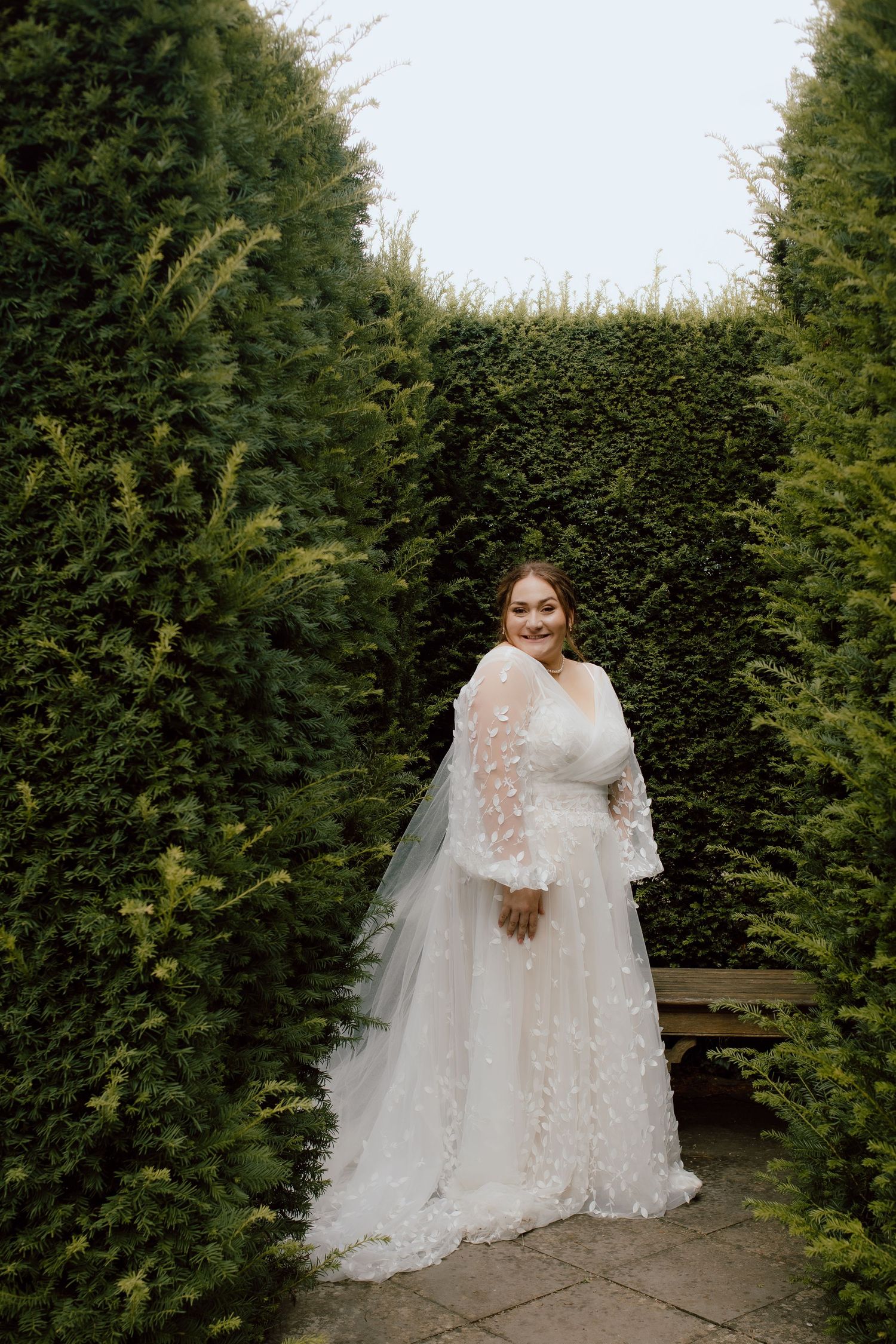 A bride in a long-sleeved lace wedding gown poses between manicured garden hedges.