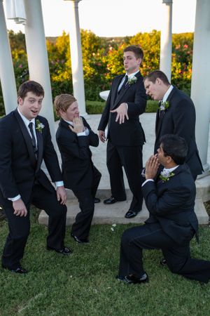Groomsmen in black suits pose playfully for photos in an outdoor gazebo at sunset.