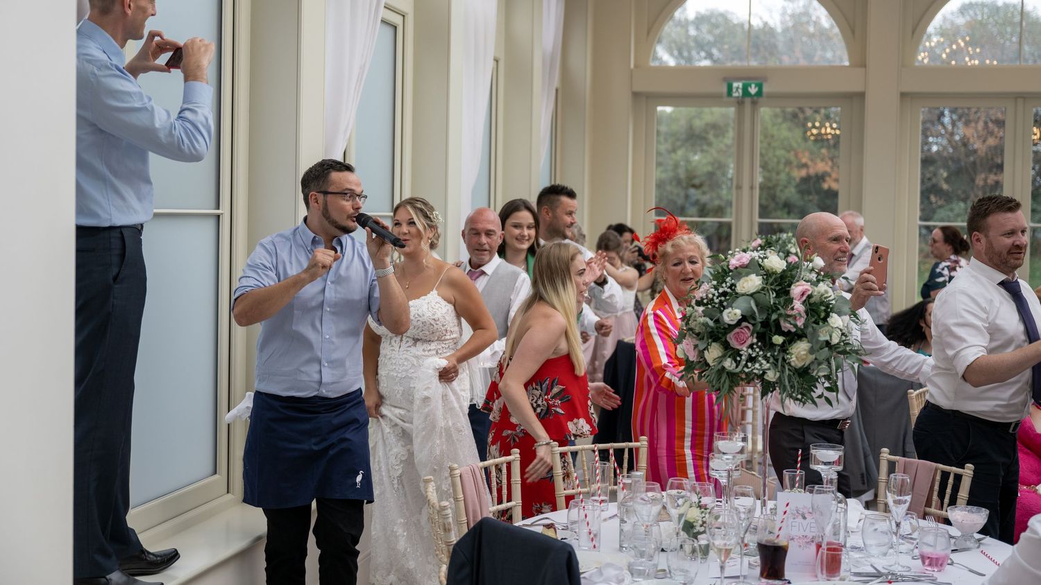 Wedding party and guests participating in group activity during reception in bright conservatory setting.