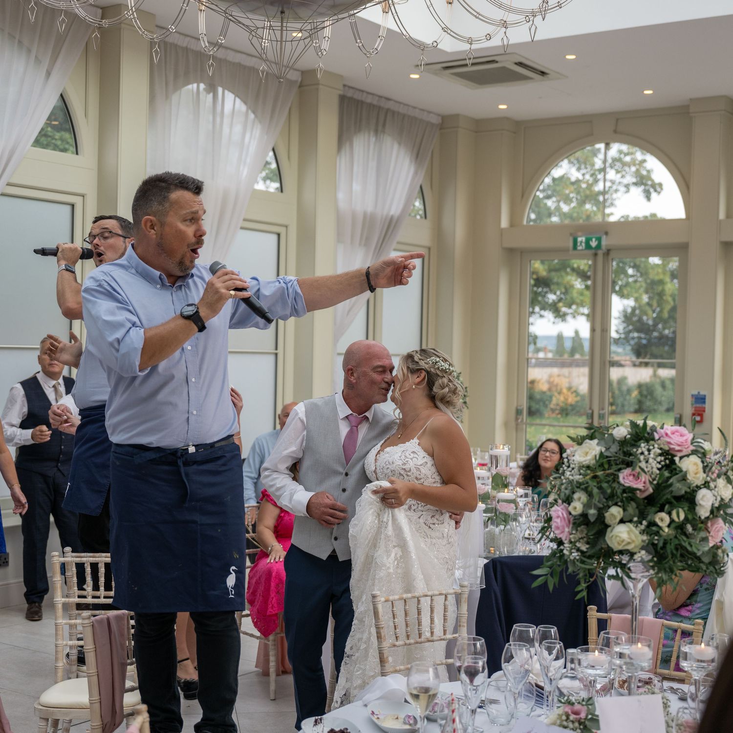 Wedding guests gathered in an elegant reception venue with chandeliers and floral centerpieces during a speech moment.