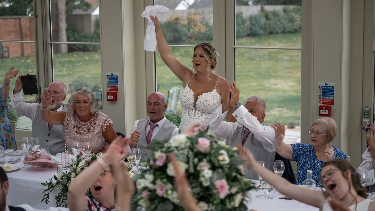 Celebratory moment at wedding reception as guests wave napkins in bright conservatory setting.