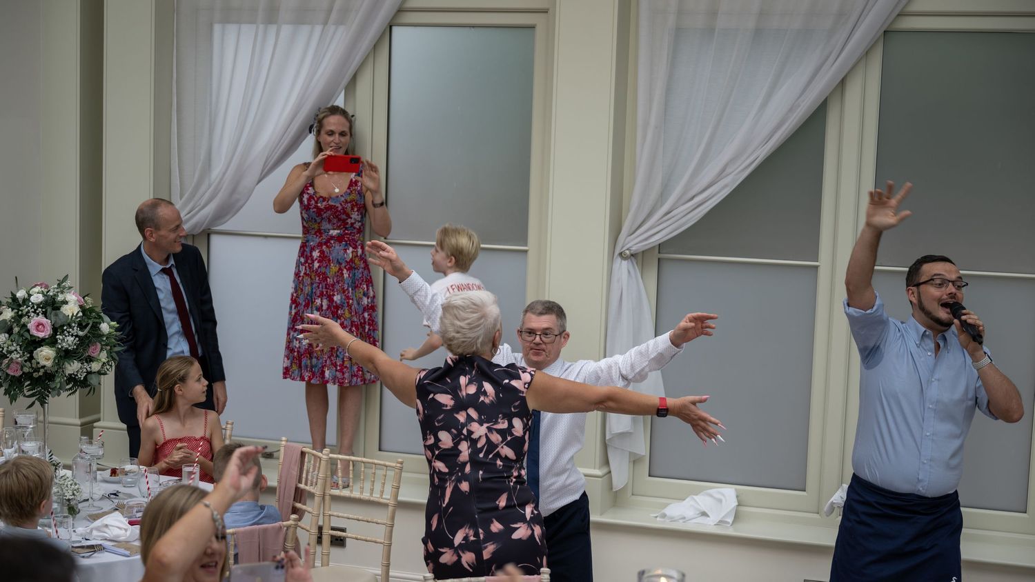 Enthusiastic wedding guests dancing and celebrating with arms raised in elegant reception venue.