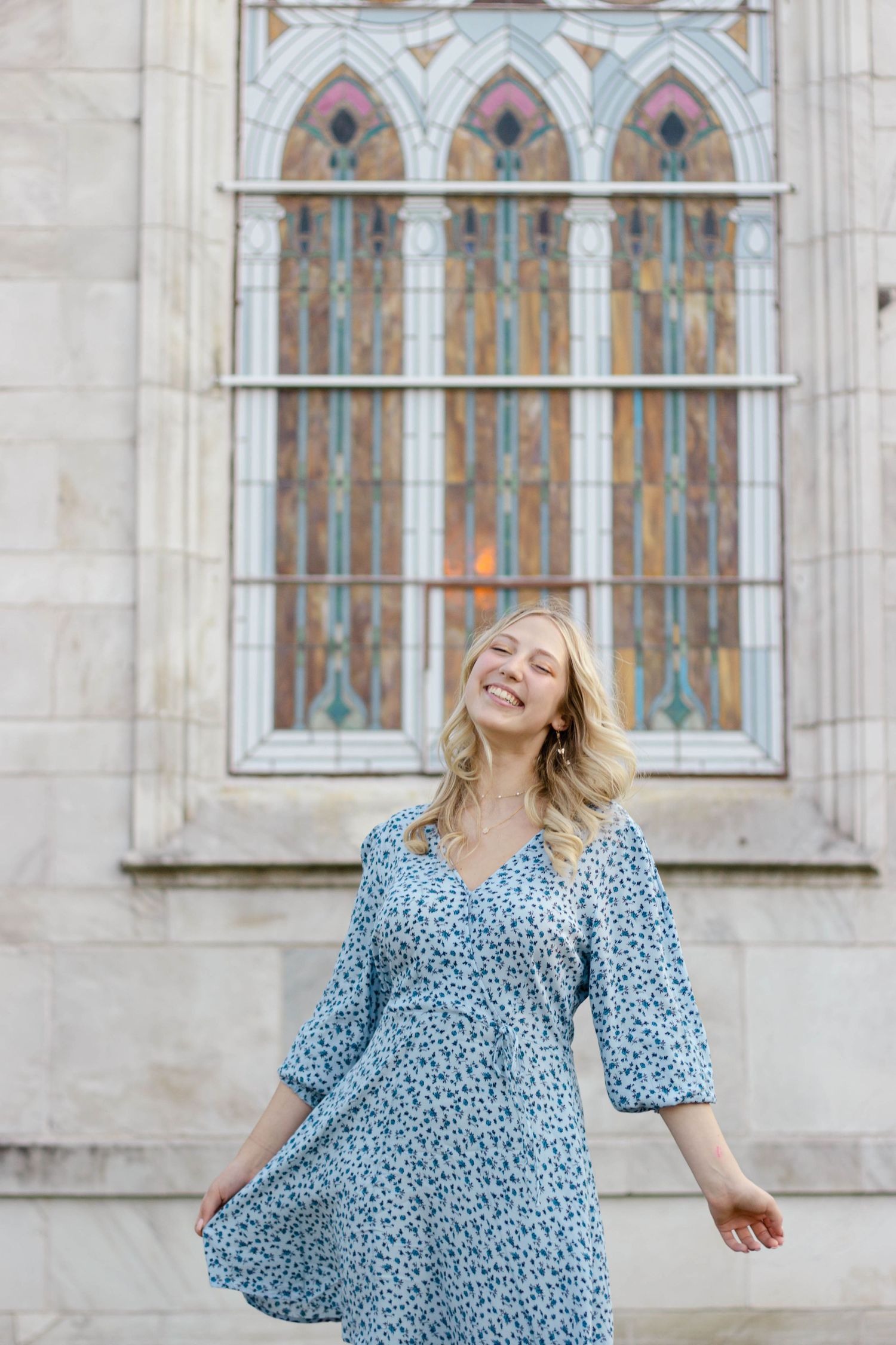 Sioux City, Iowa photographer. Smiling blonde woman in blue floral dress posing before stained glass church window.