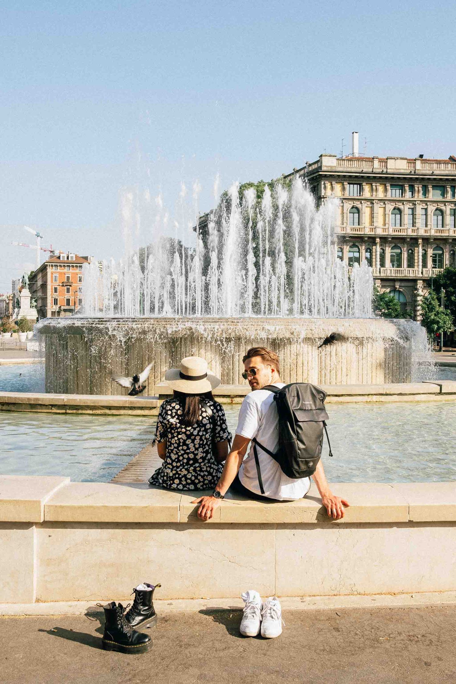 Two people sit on edge of fountain in Castello square in Milan with backpack and shoes on ground, water spraying in background.