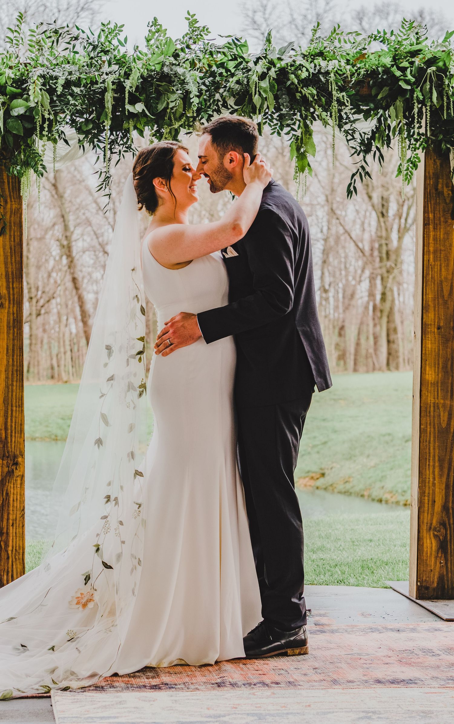 Newlyweds share intimate moment under greenery arch during outdoor wedding ceremony.