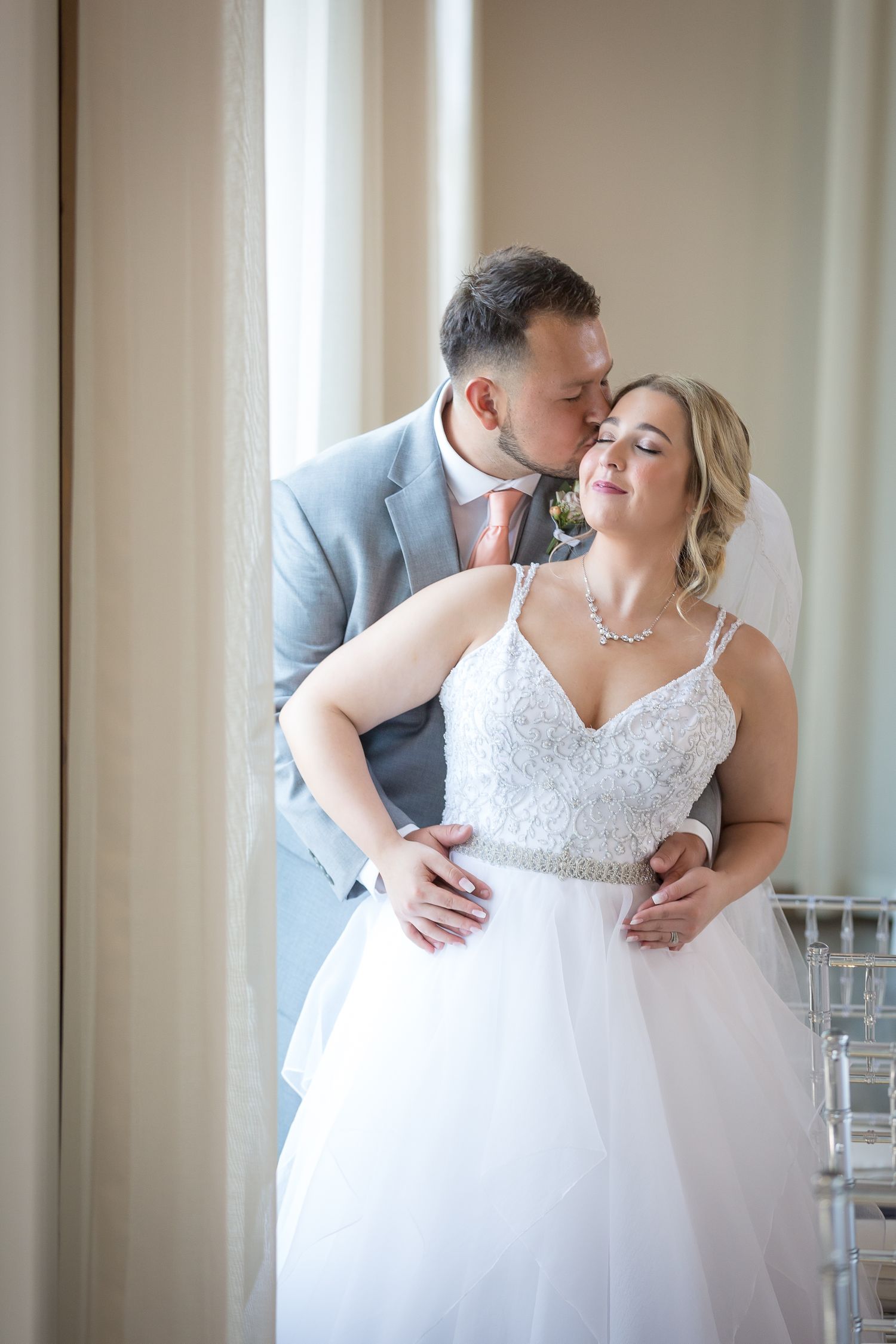 groom standing behind bride, kissing her on the cheek, near window of texas wedding venue