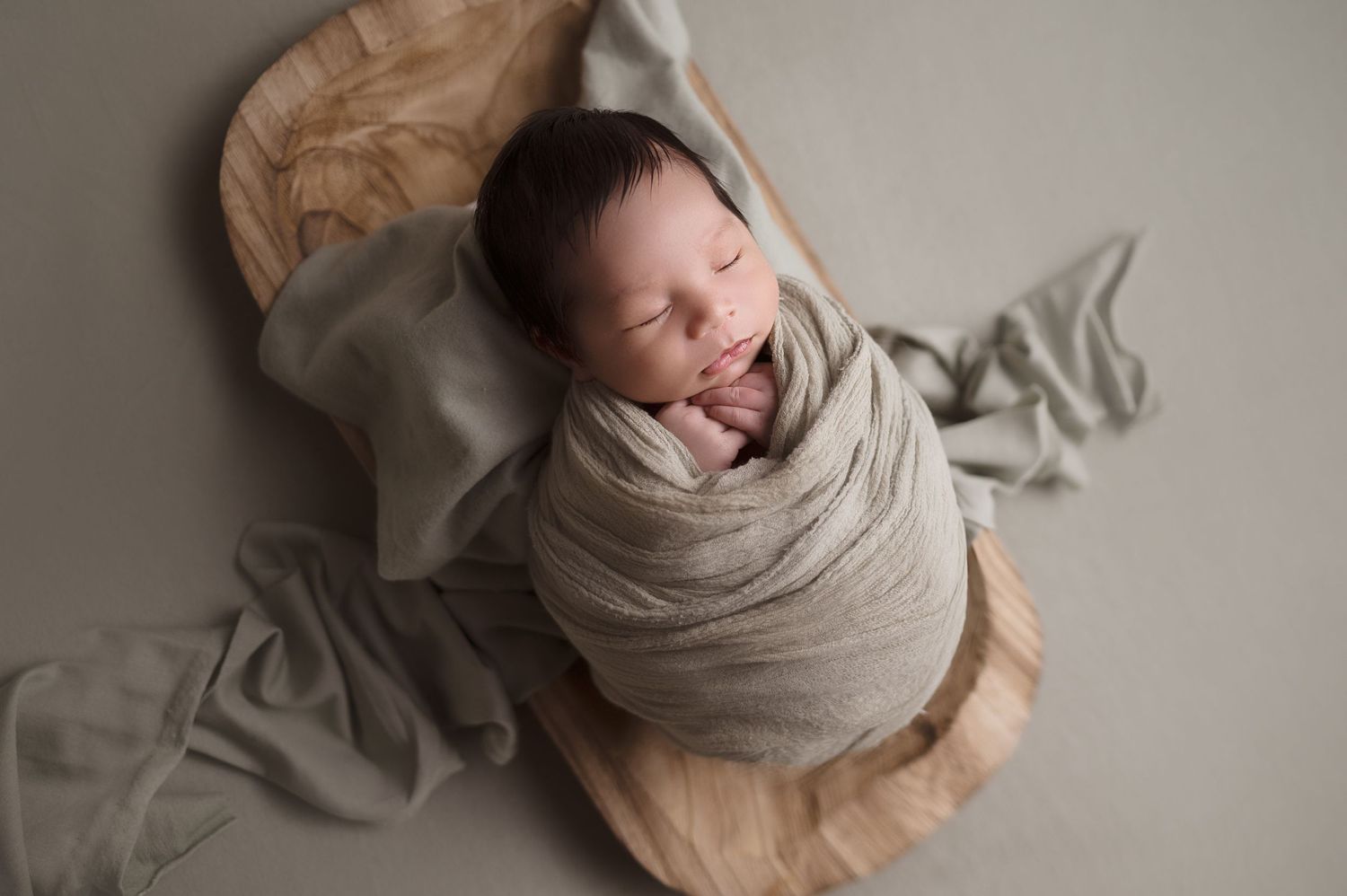 Sleeping baby wrapped in neutral swaddle on textured beige surface.
