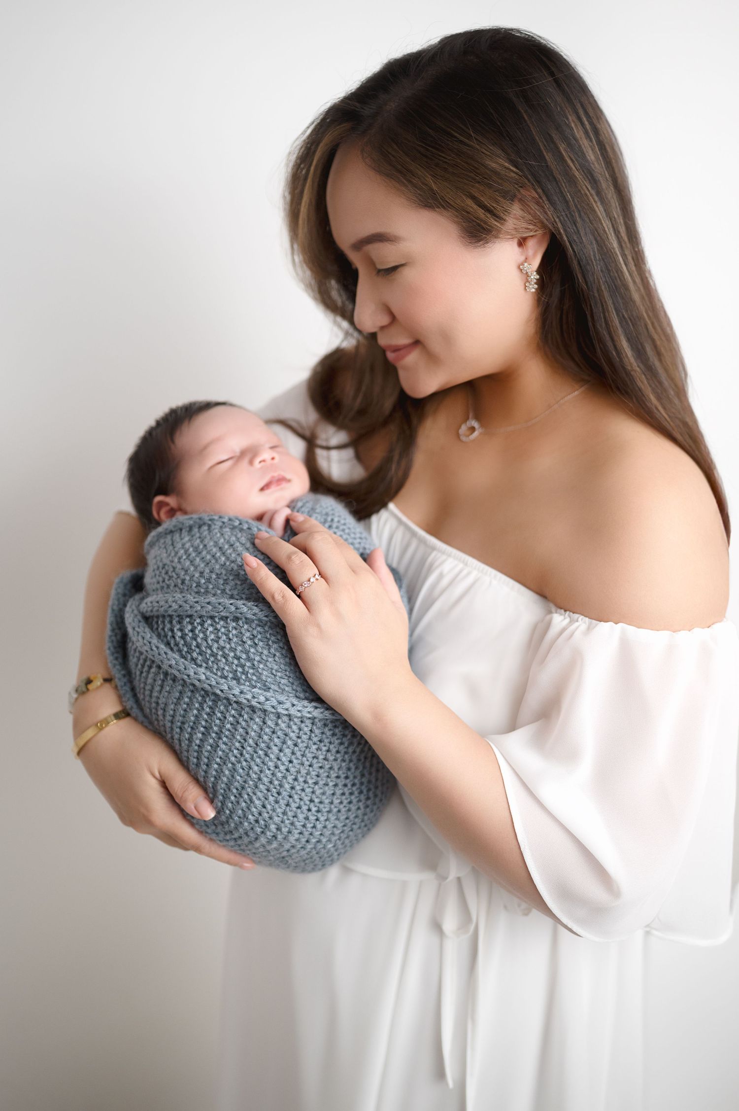 Mother in white blouse holding baby wrapped in gray knit blanket.
