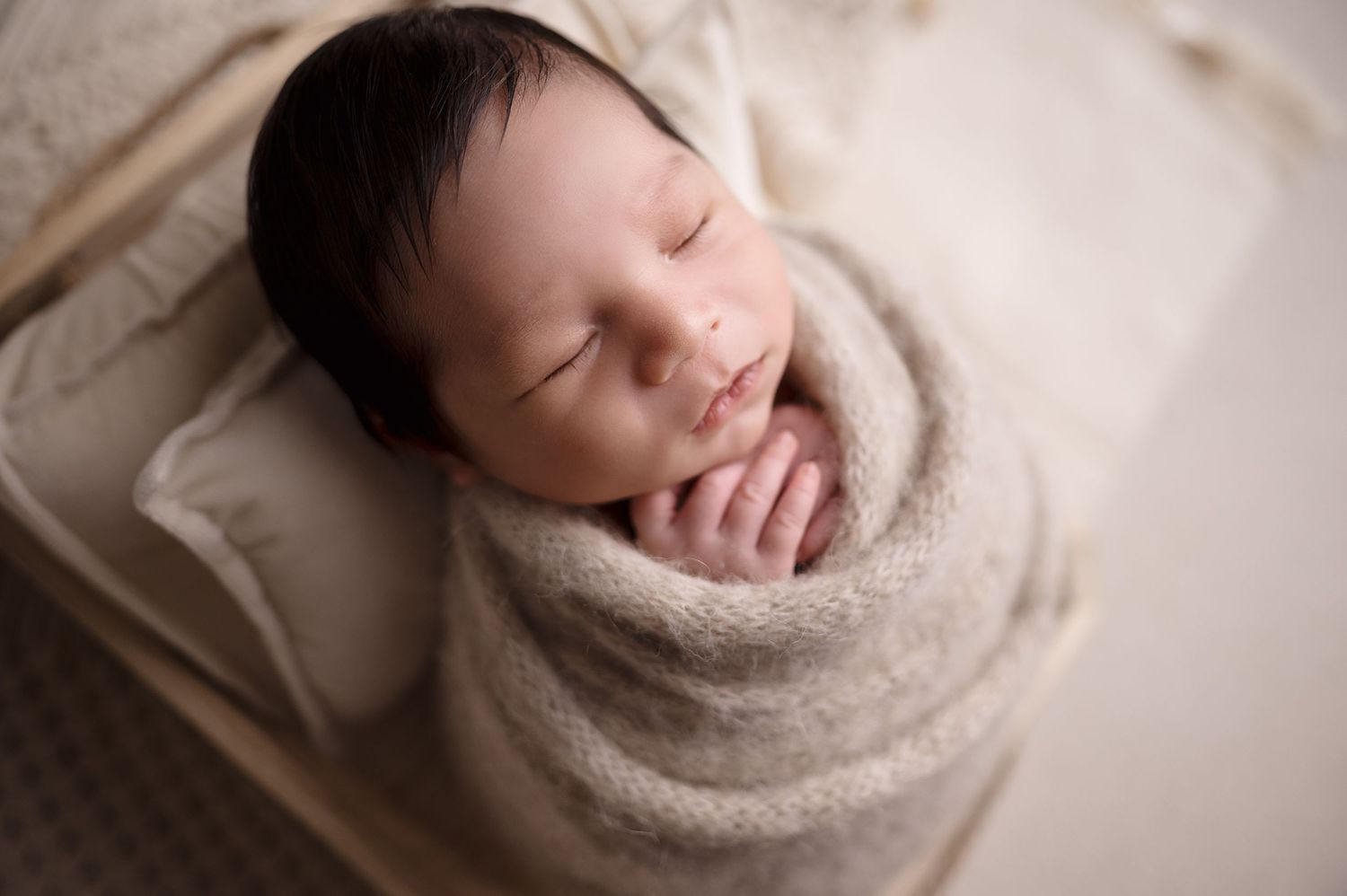 Sleeping newborn wrapped in soft cream colored fabric on light background.