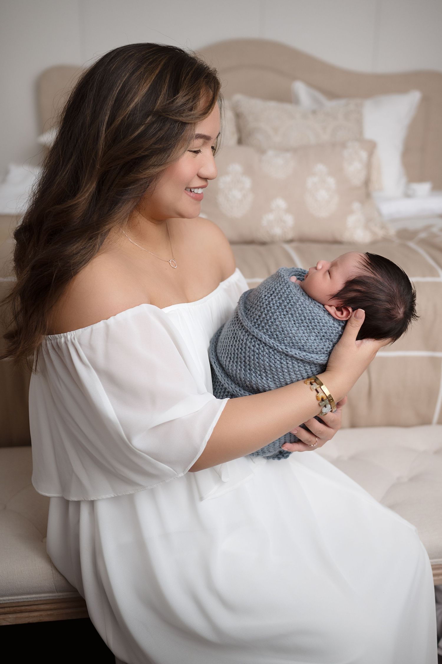 Mother in flowing white dress cradles newborn on beige bedding.