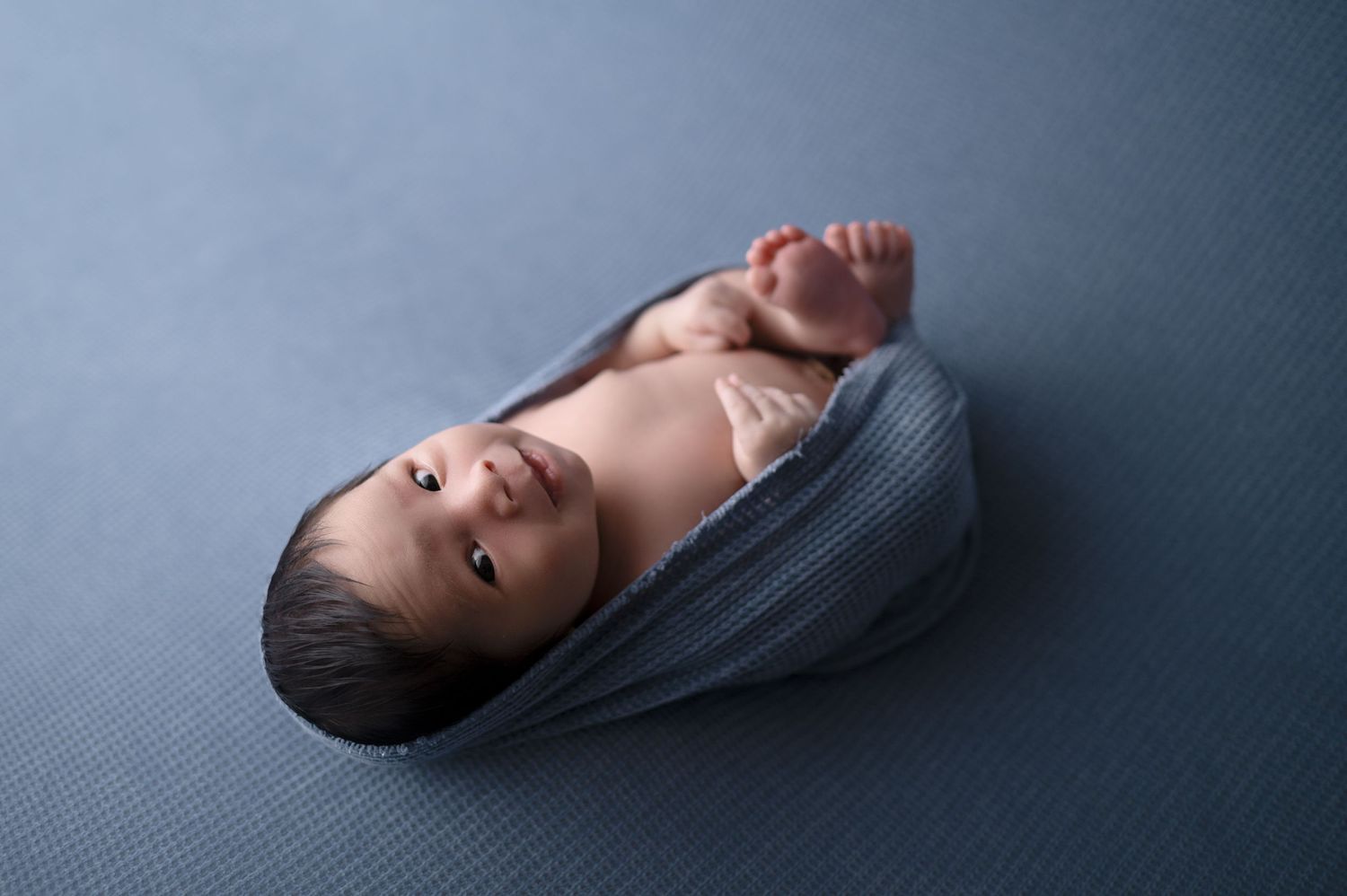 Sleeping newborn wrapped in dark fabric against blue backdrop.