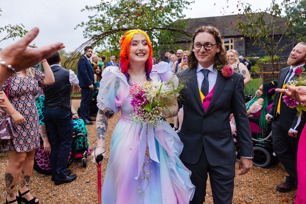 Eliza and Michael walking through their confetti arch at their wedding in their rainbow wedding clothing smiling