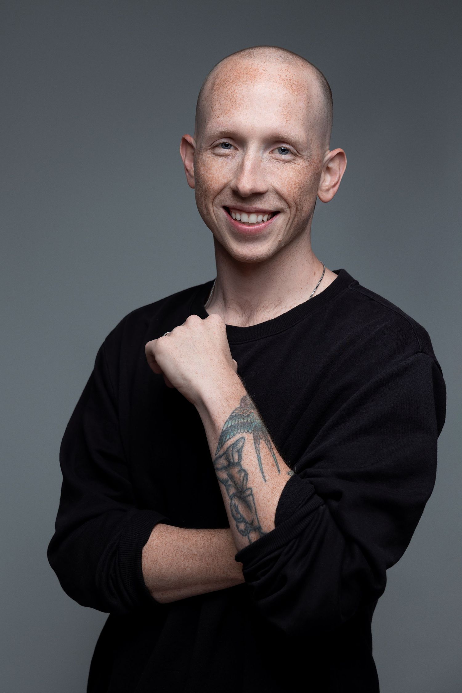 Studio portrait of person in black sweater showing forearm tattoo against gray backdrop.