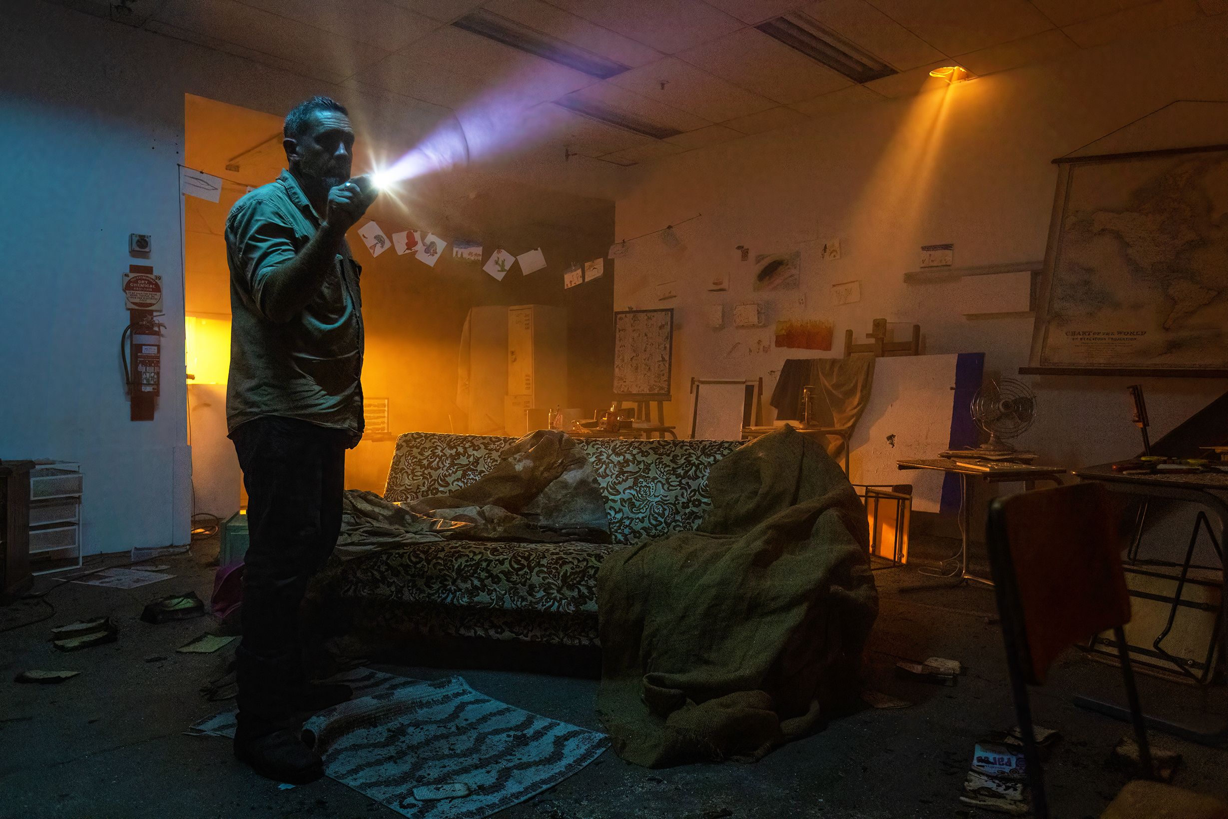 A man stands in a dark room shining a flashlight into the darkness. The room is filled with haze and lit by orange light