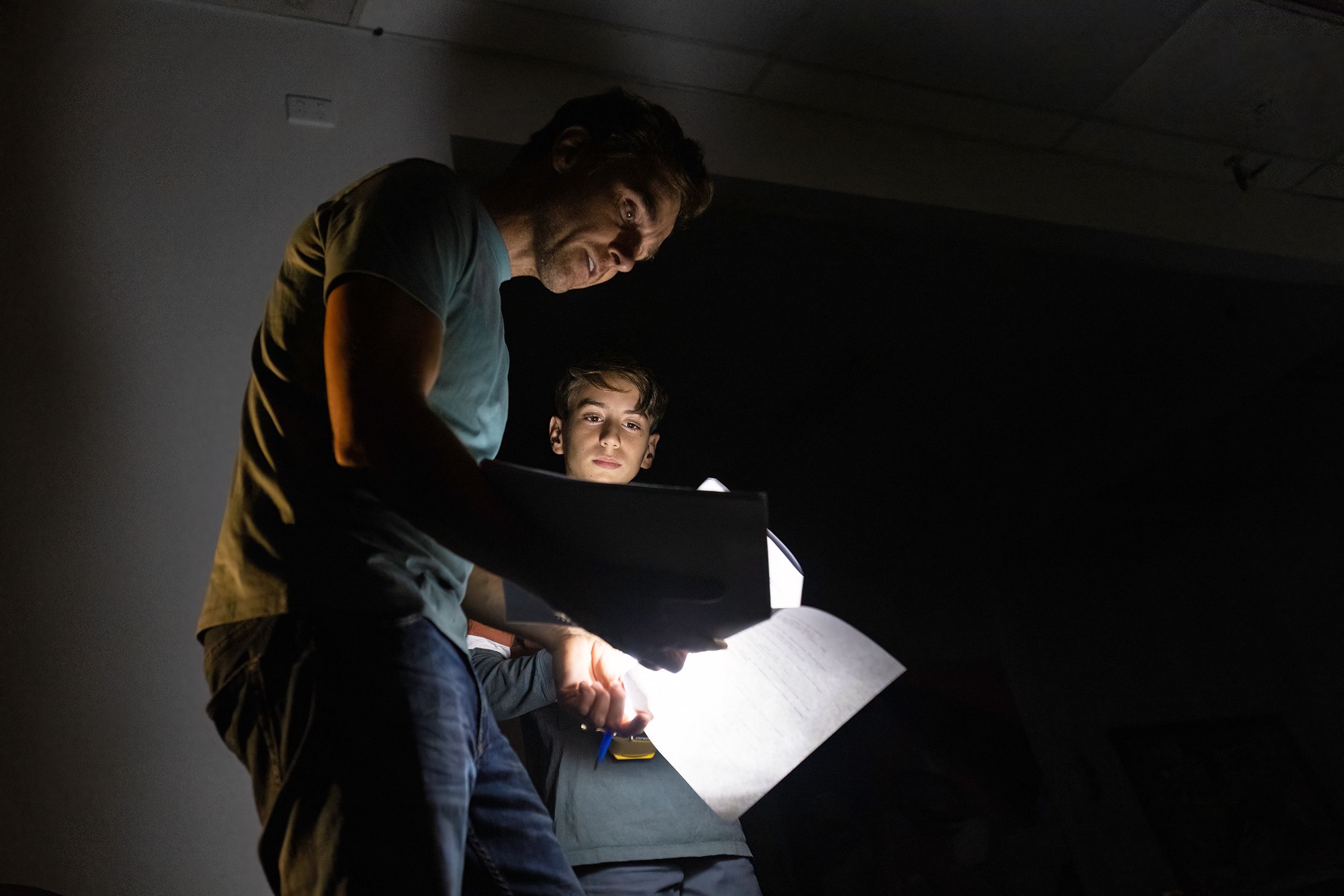A film director looks over a script lit by a flashlight with a young actor on a very dark filmset.