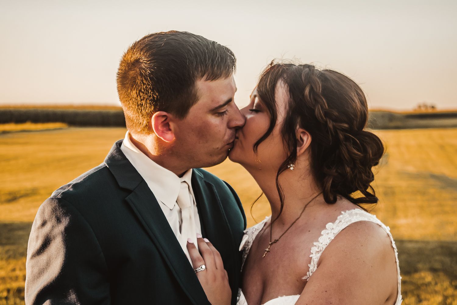 Romantic sunset kiss between newlyweds in formal attire against golden field backdrop.