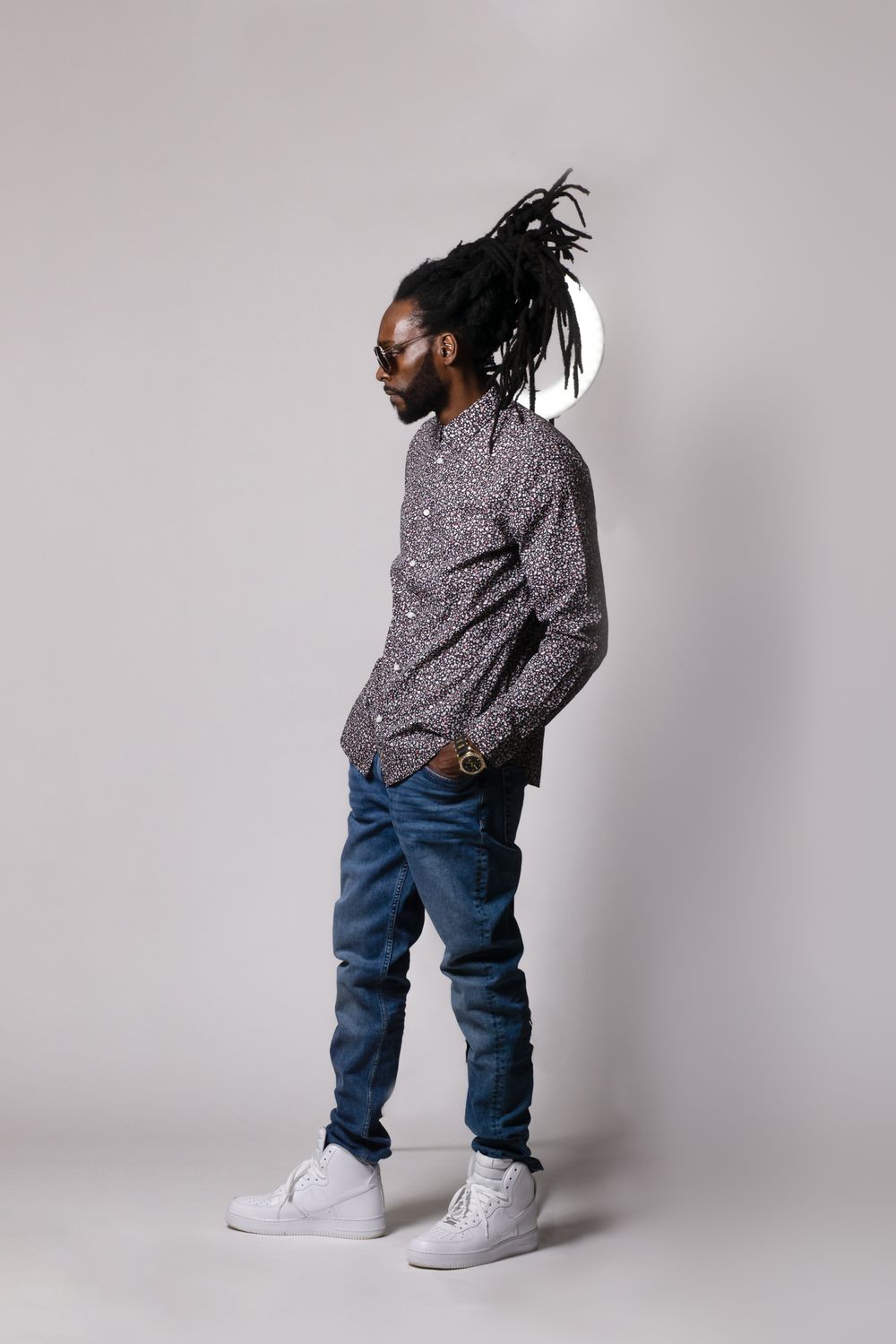 Model in casual wear with dreadlocks poses against grey background wearing patterned shirt and jeans.