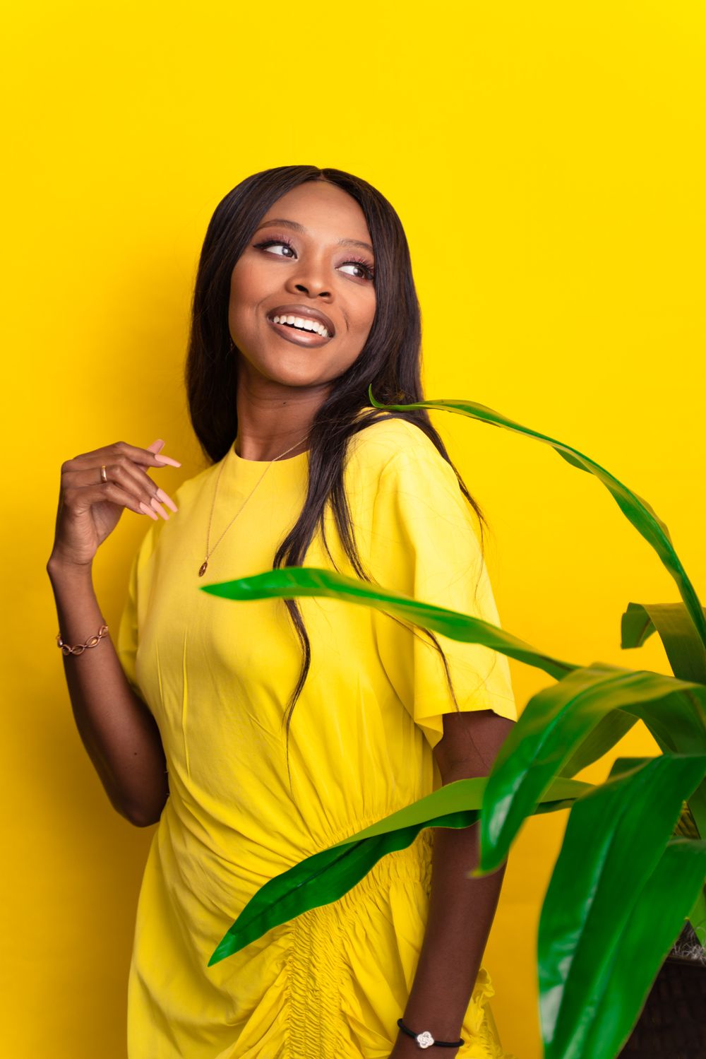 Woman in yellow dress poses against bright yellow background with tropical plant leaves.