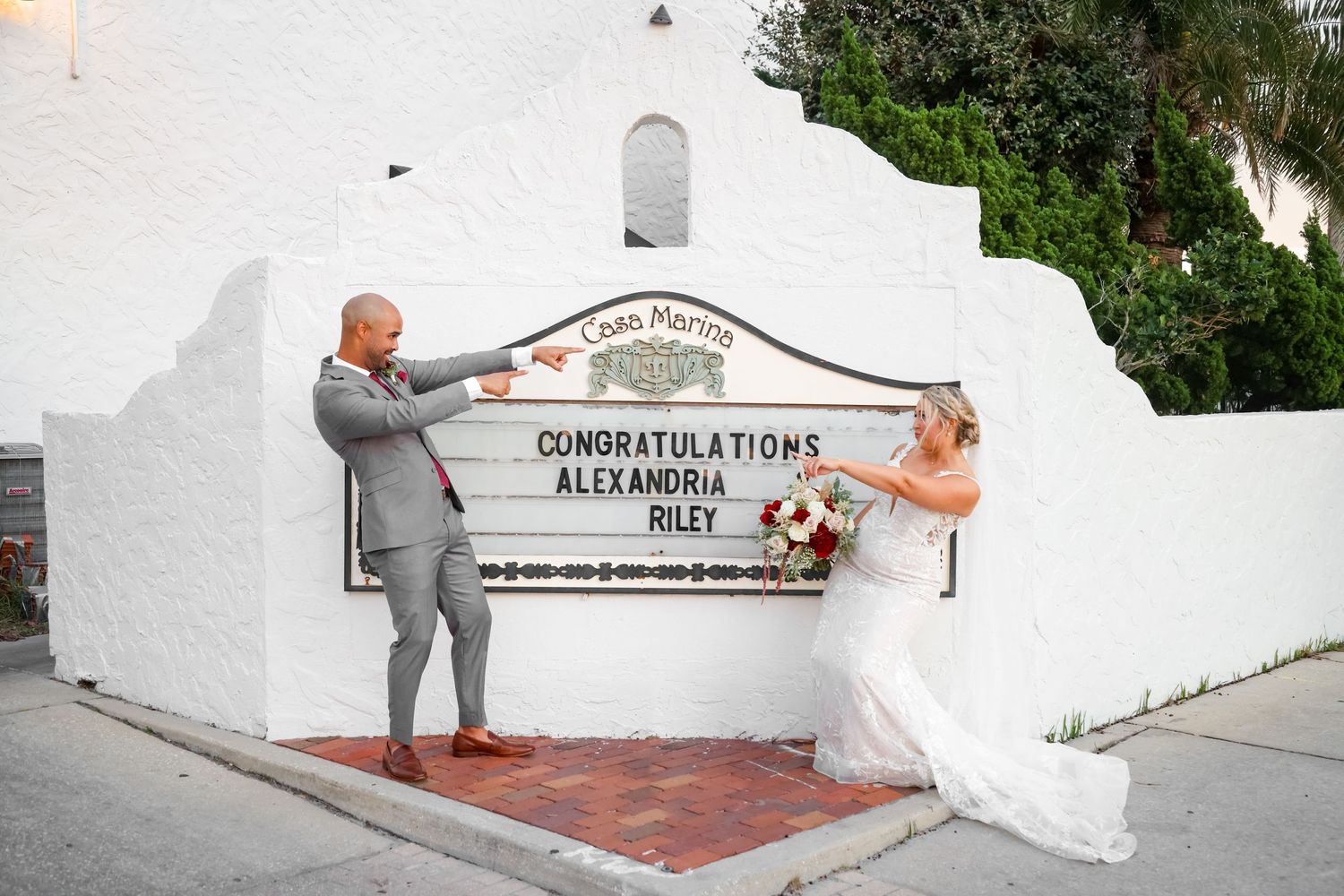 A wedding couple poses in front of a white Spanish-style church wall with congratulatory text.