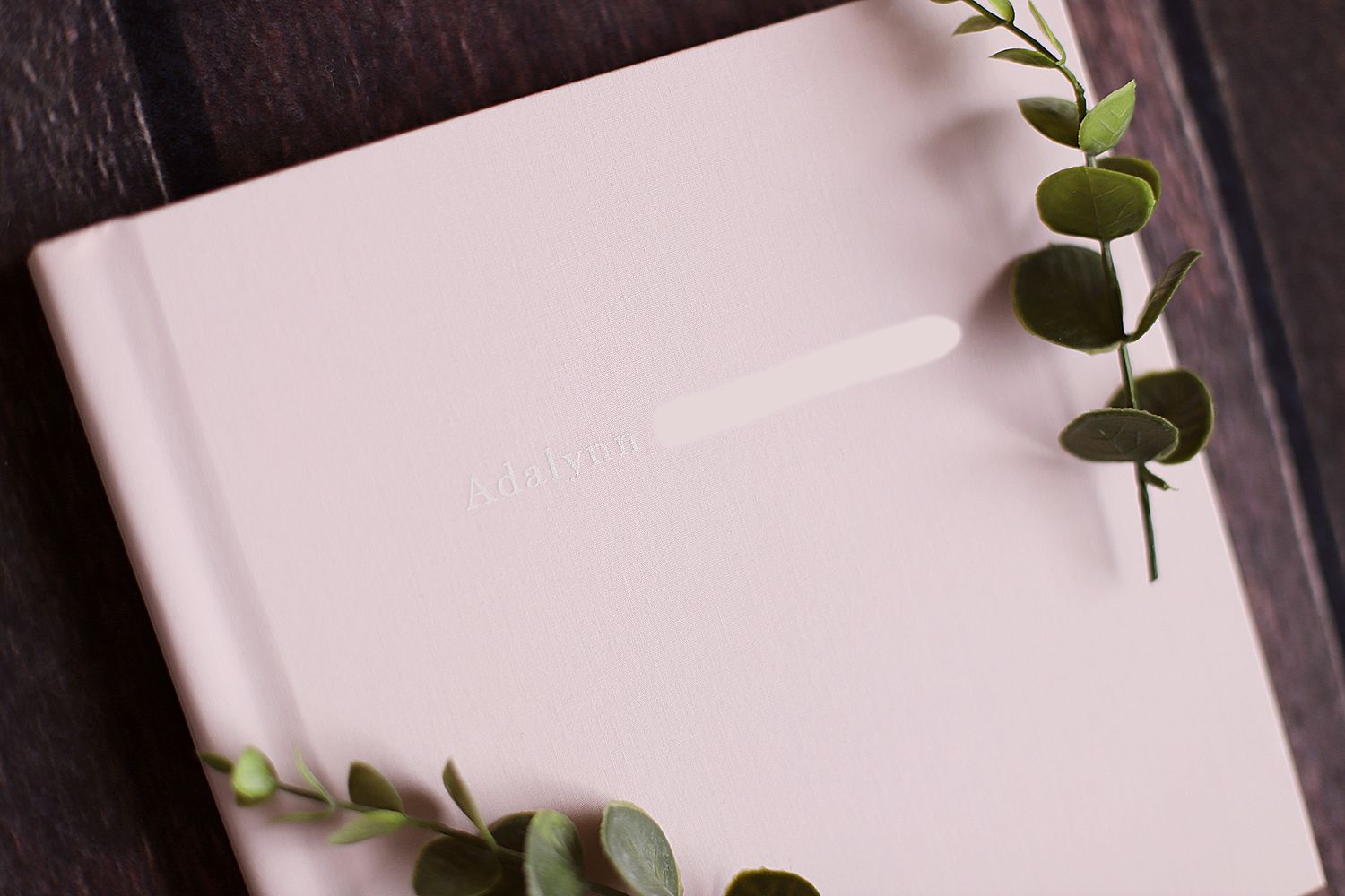 Pink photo album cover adorned with fresh eucalyptus leaves on dark wooden background.