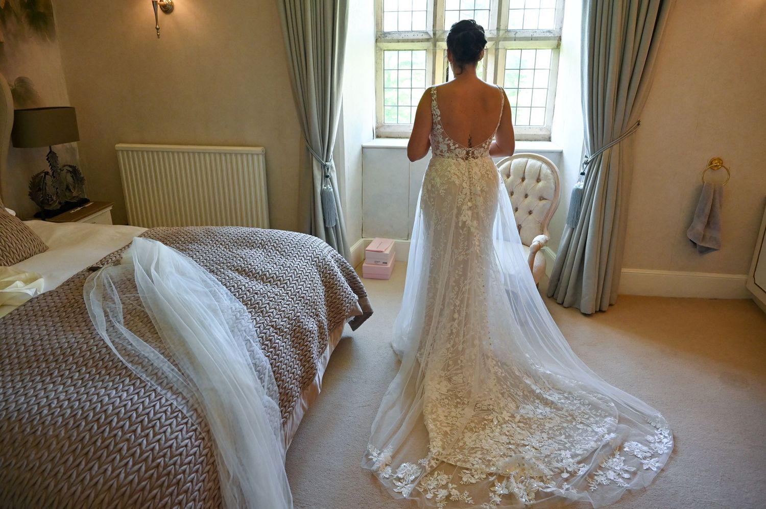 Bride in lace wedding gown with dramatic train stands by window in elegant bedroom.