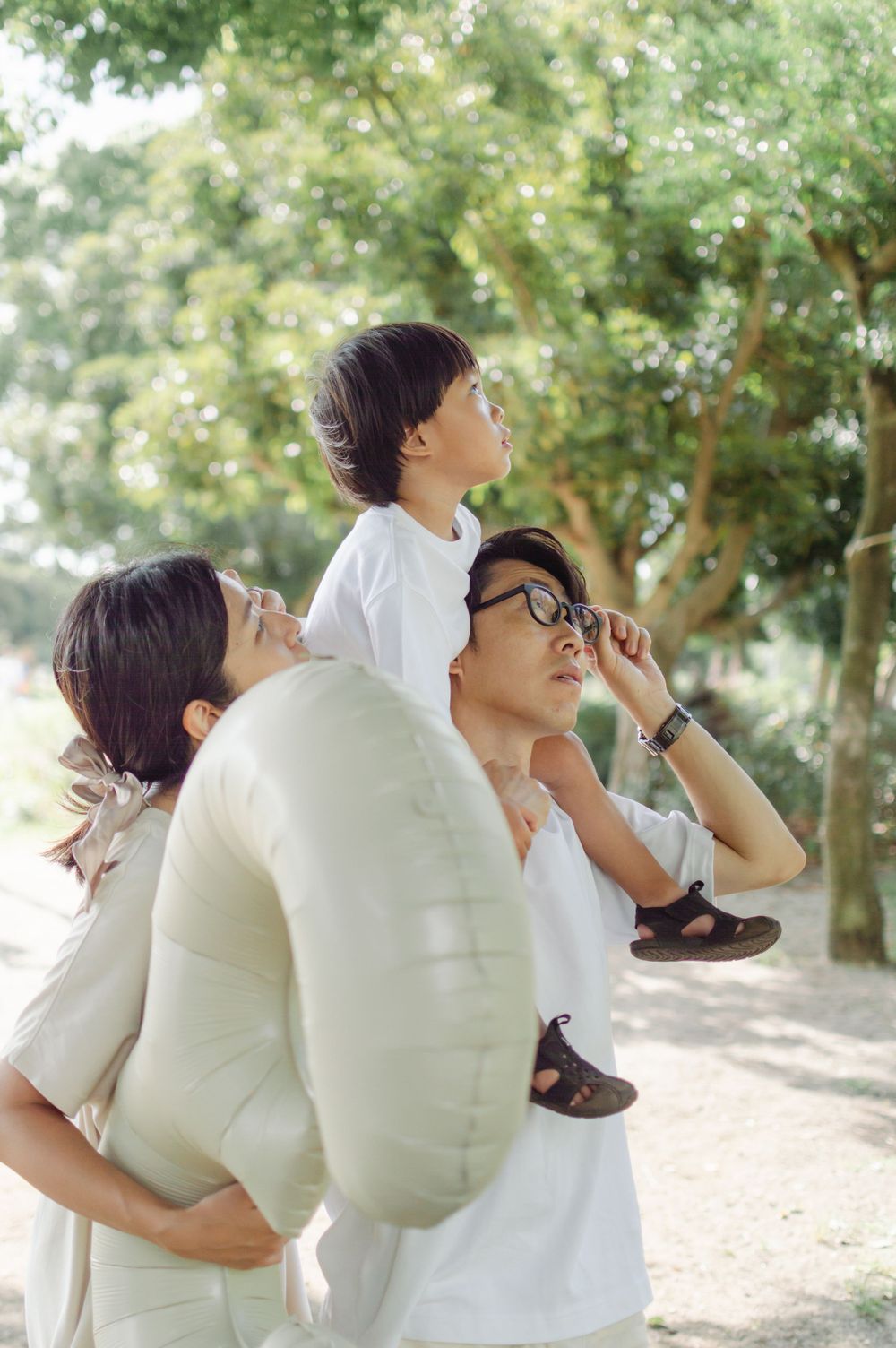 Asian family with boy on fathers shoulders and mother in sunny green park.