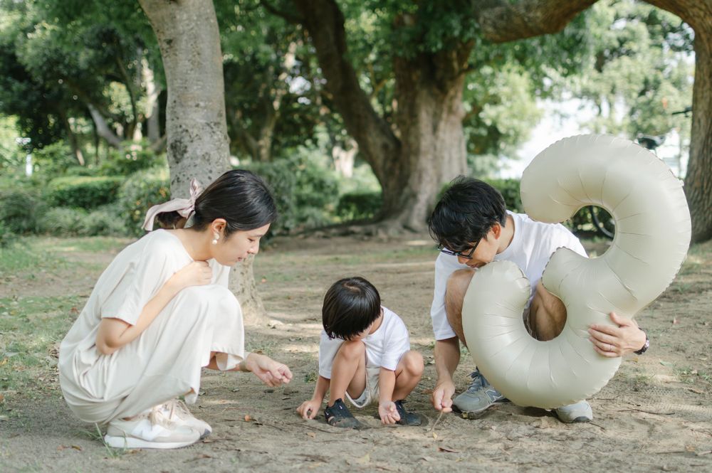 Asian family crouching together exploring the ground under large trees in a park.