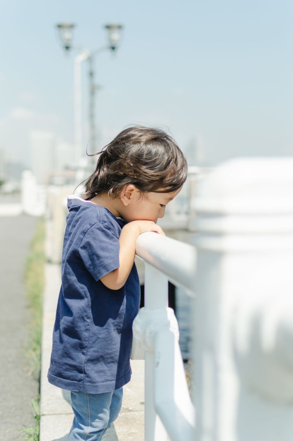 Curious young Asian child leaning over a white railing outdoors on a sunny day.