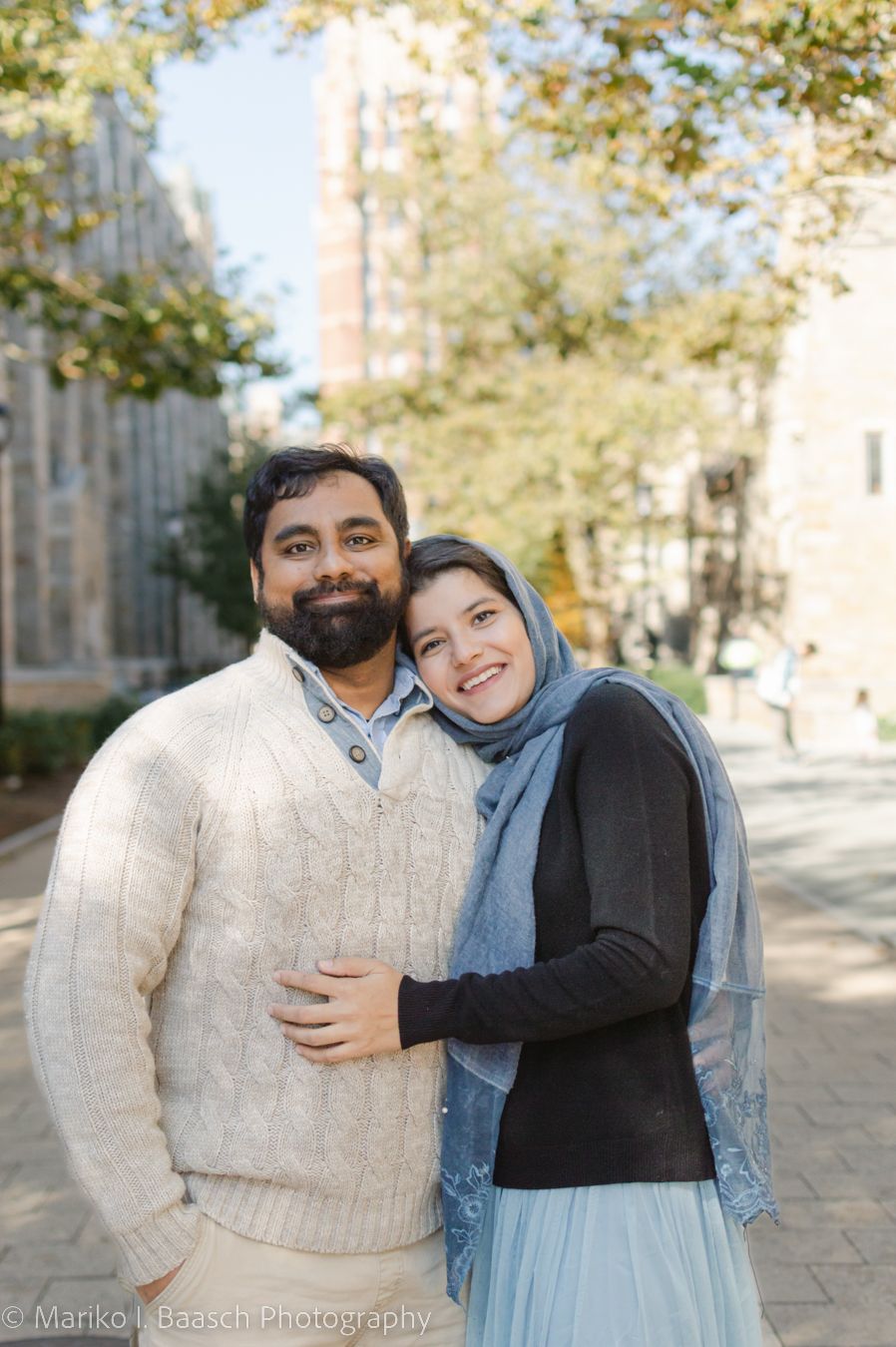 A smiling couple poses together on a sunny campus pathway with gothic architecture.